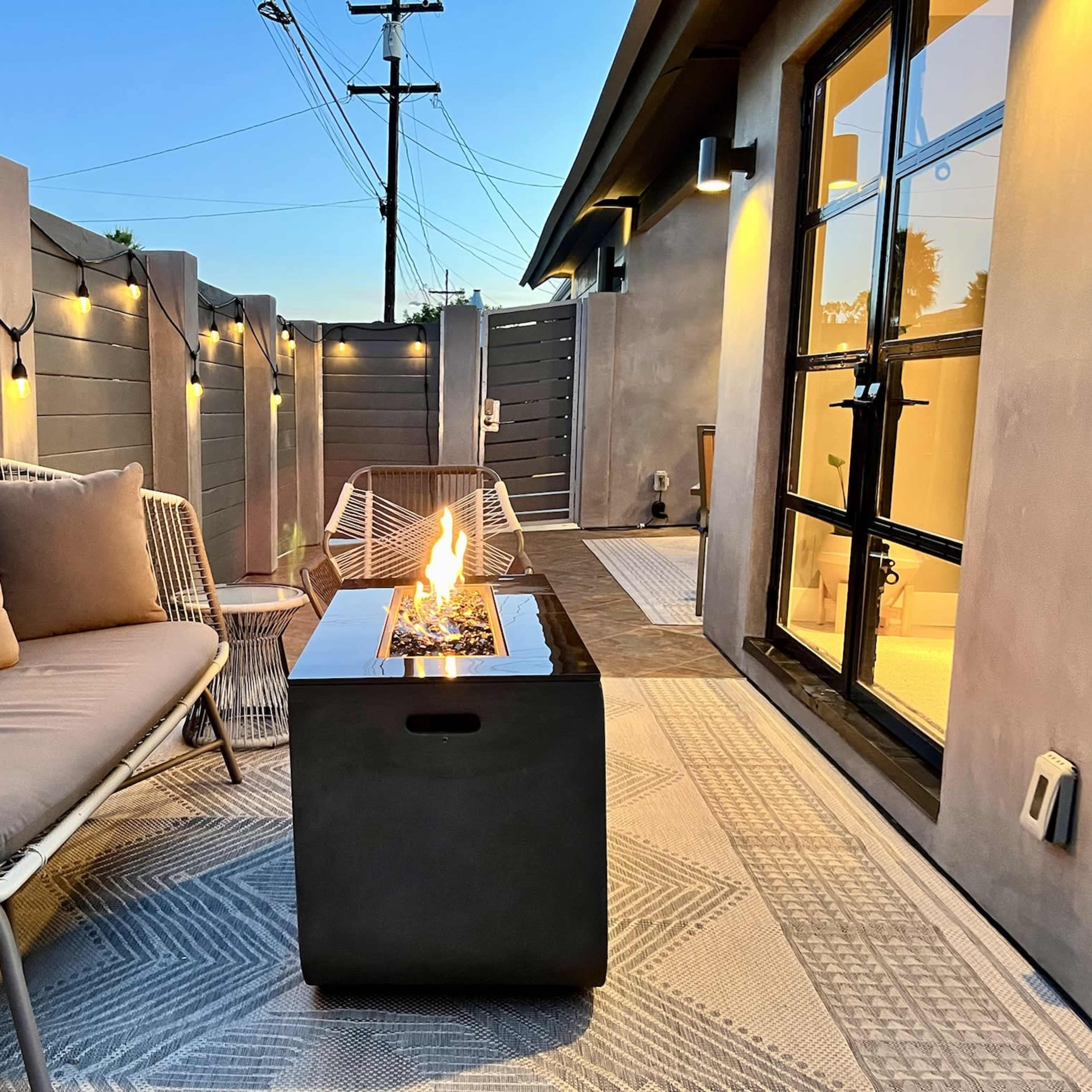 A modern outdoor seating area features a fire pit table surrounded by woven chairs and illuminated by string lights.