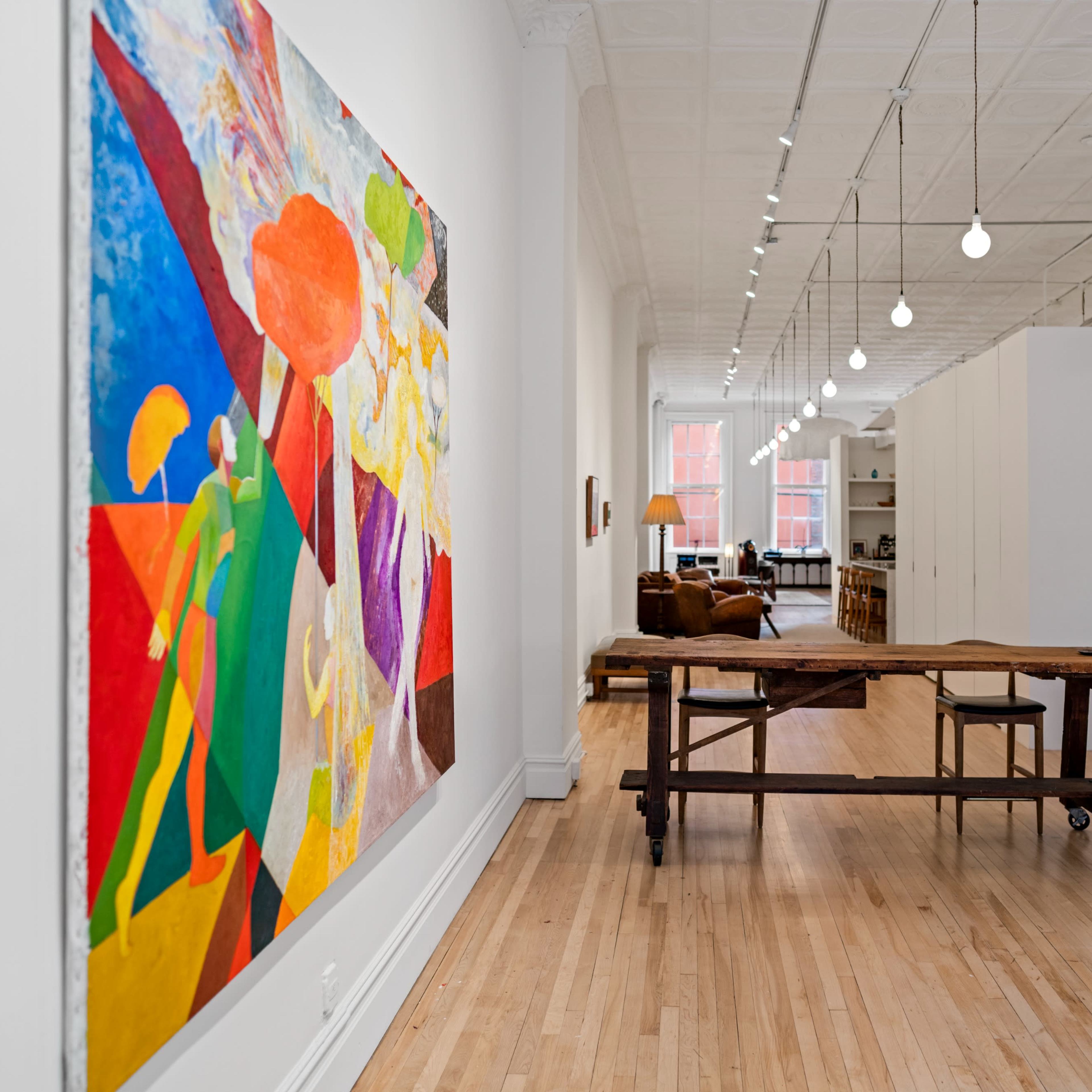 A brightly lit gallery space featuring a large colorful painting on the left wall and a wooden table with two stools in the center, leading to a cozy living area in the background.