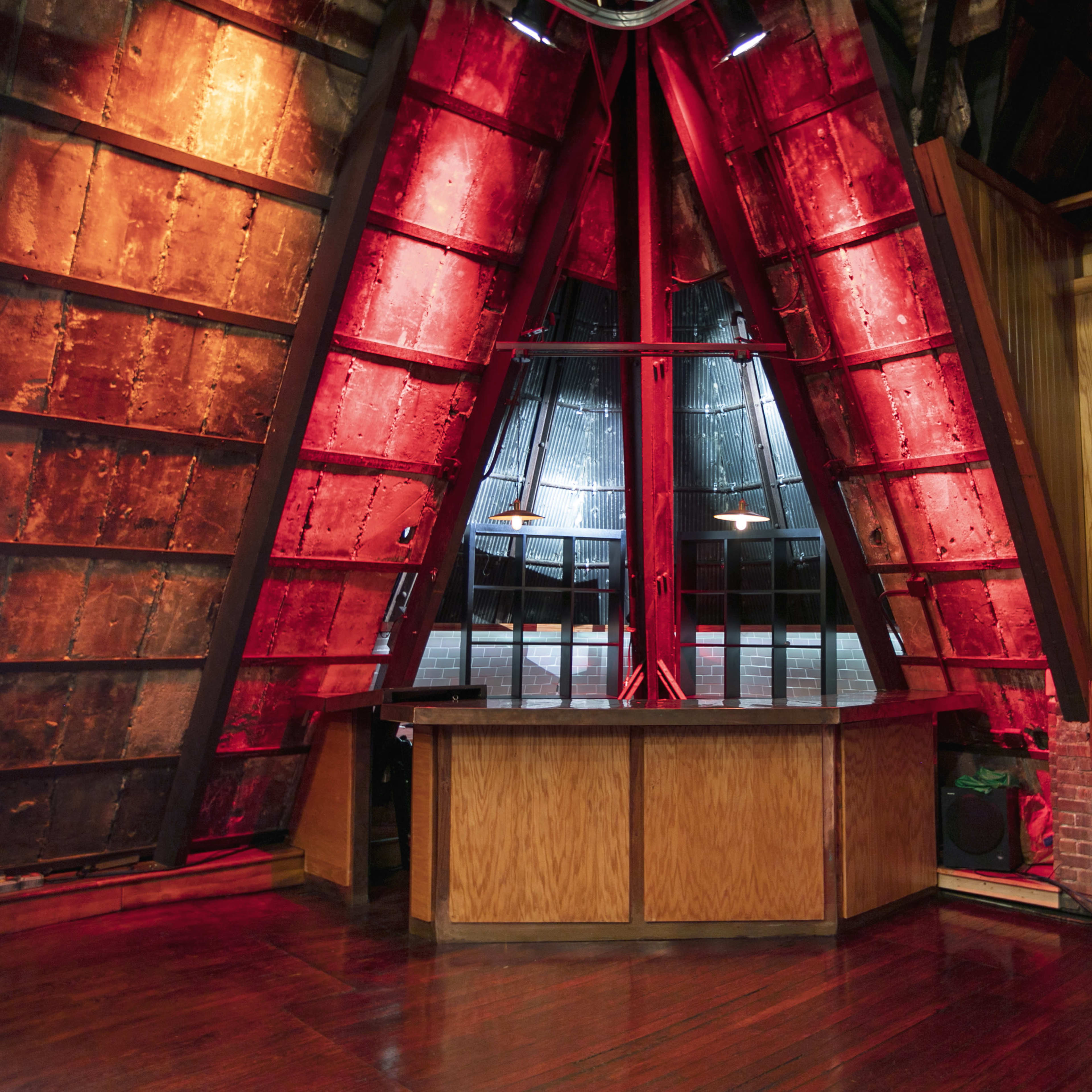 A triangular wooden bar in a dimly lit room with red and black walls, featuring large windows and industrial-style lighting.