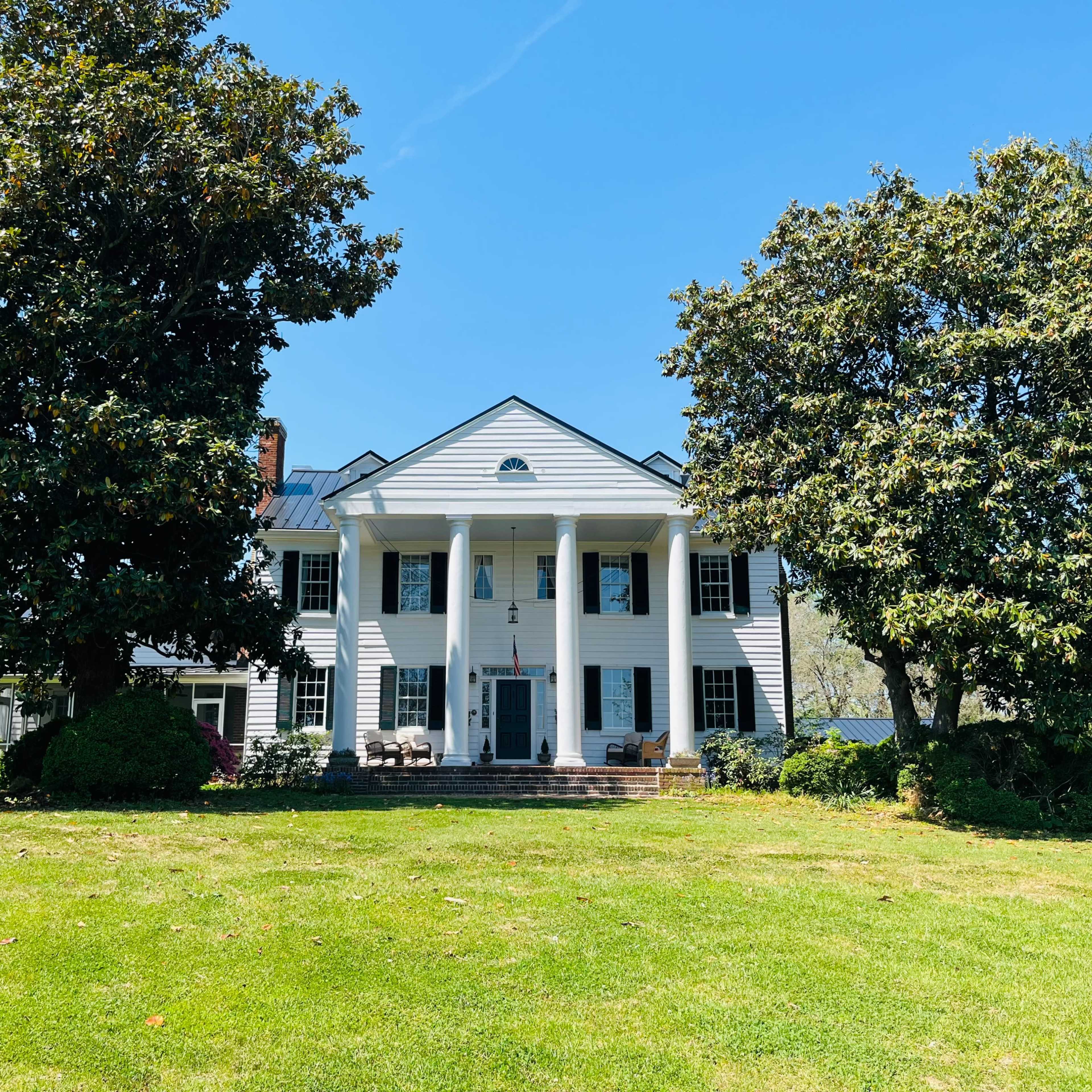 A large white colonial-style house with six columns and a manicured lawn is surrounded by tall trees under a clear blue sky.