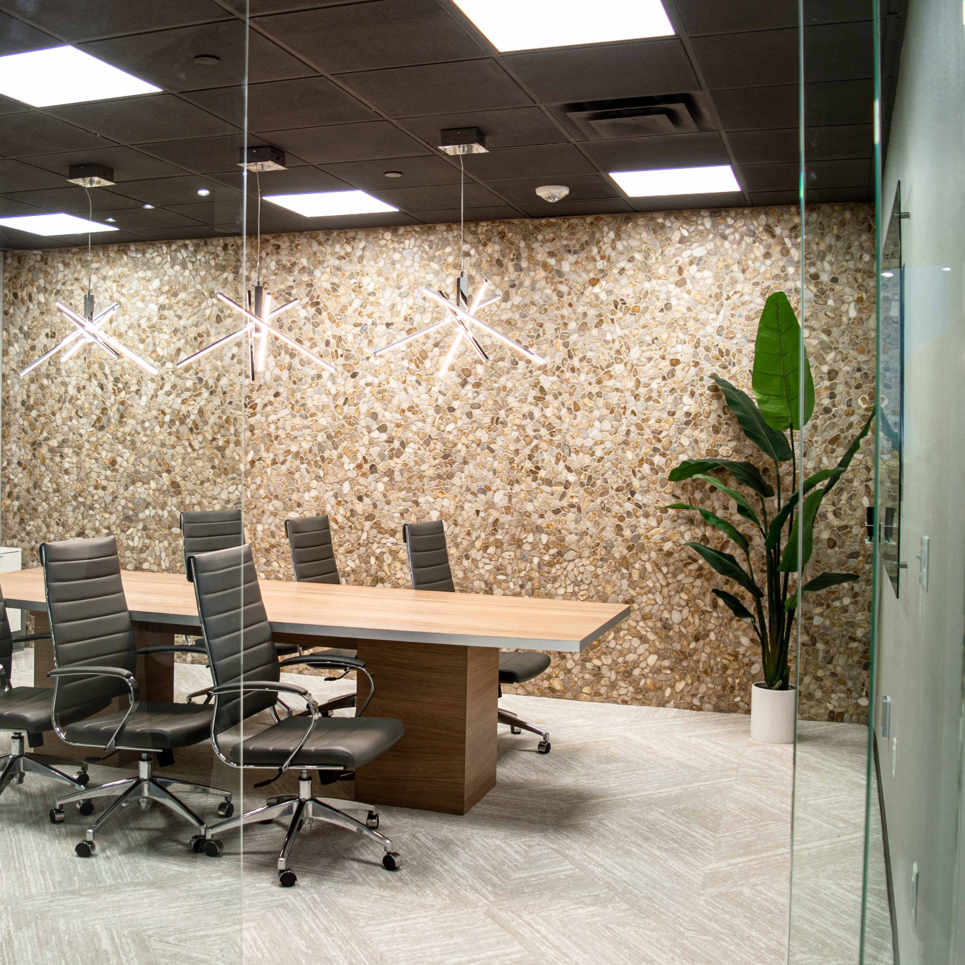 A modern conference room features a large wooden table surrounded by six ergonomic chairs, with a textured stone wall and a potted plant in the corner.