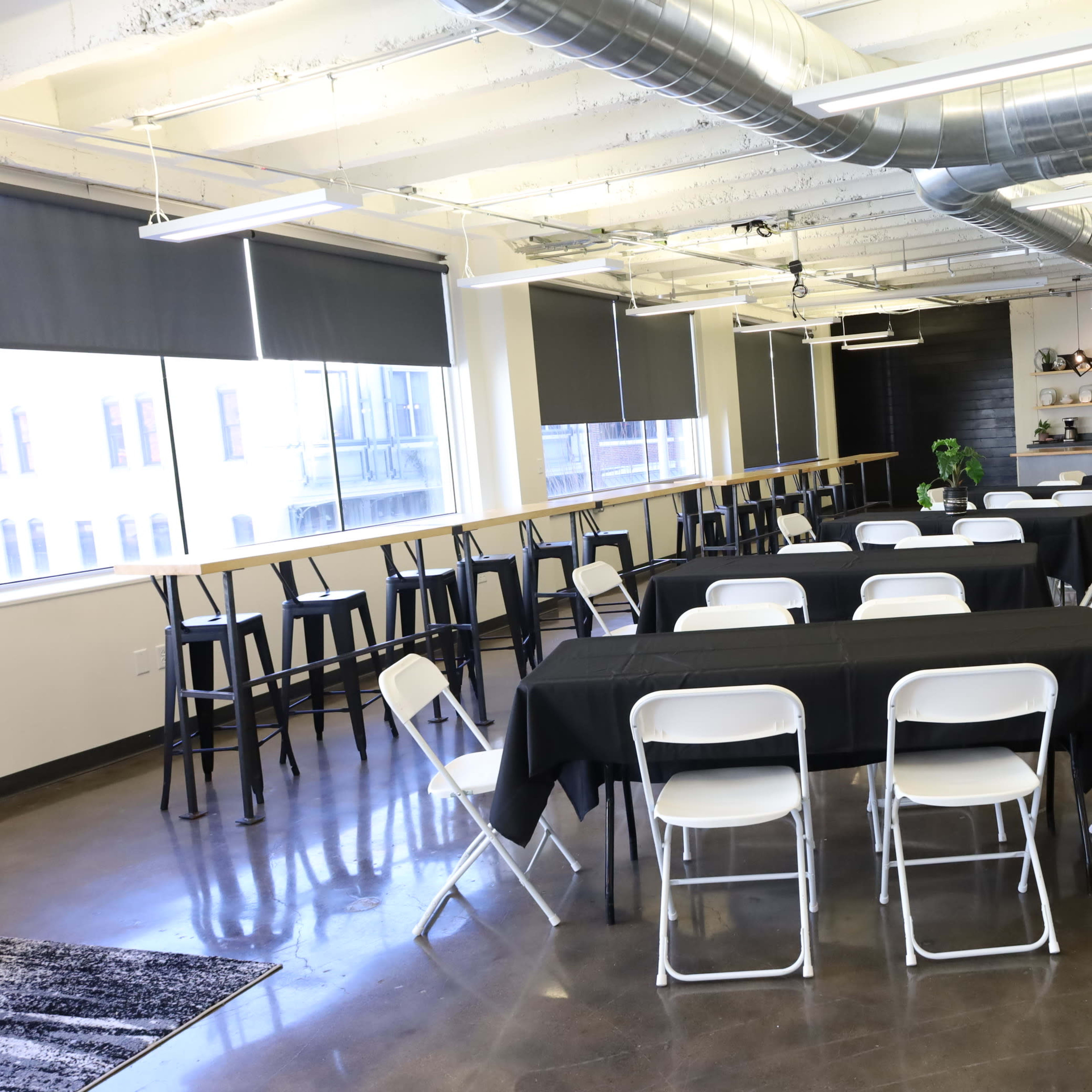 A brightly lit event space with black tables, white folding chairs, and plants, featuring large windows and a kitchenette area in the background.