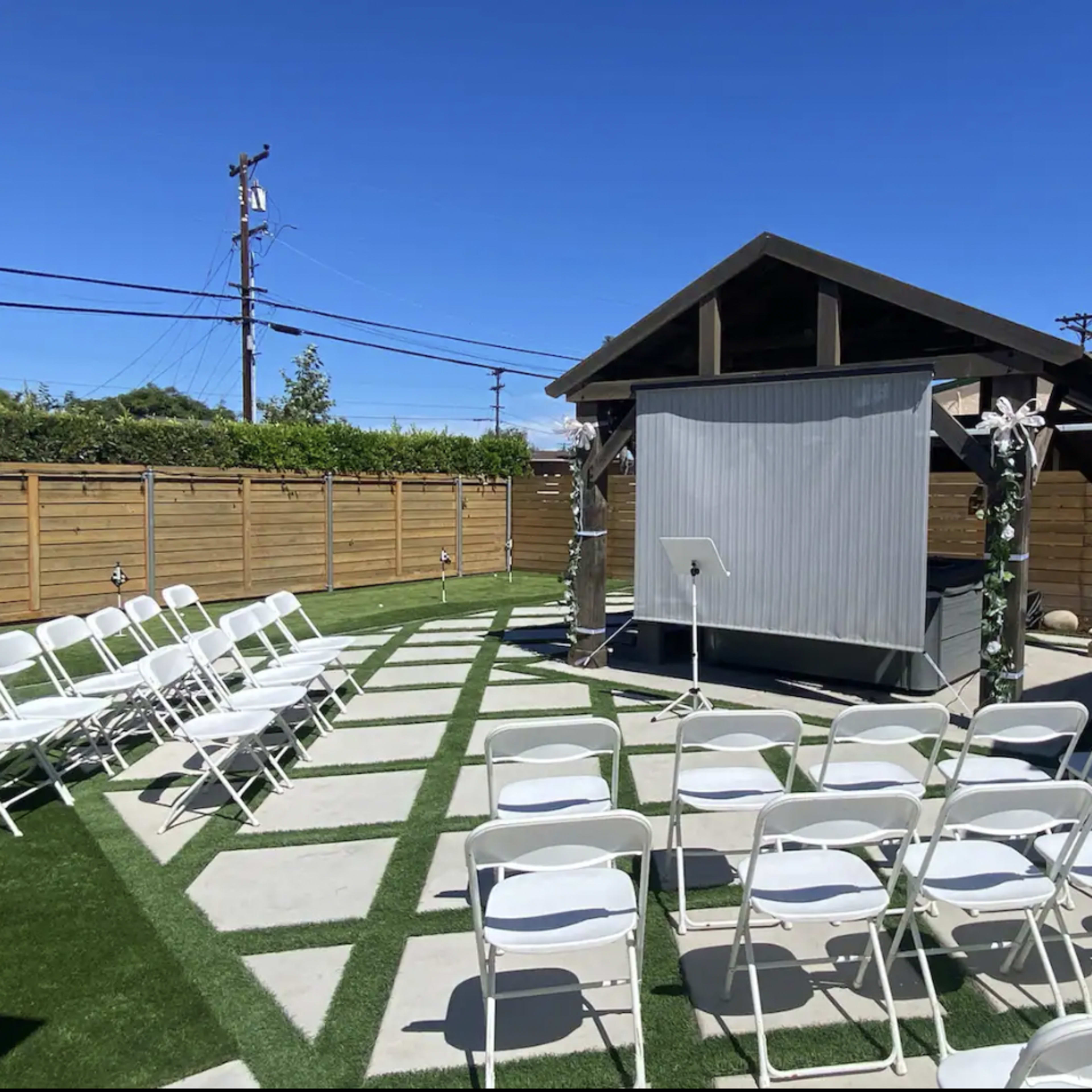 A small outdoor event space features white folding chairs arranged in front of a gazebo with a presentation screen, surrounded by landscaped grass and paving stones.