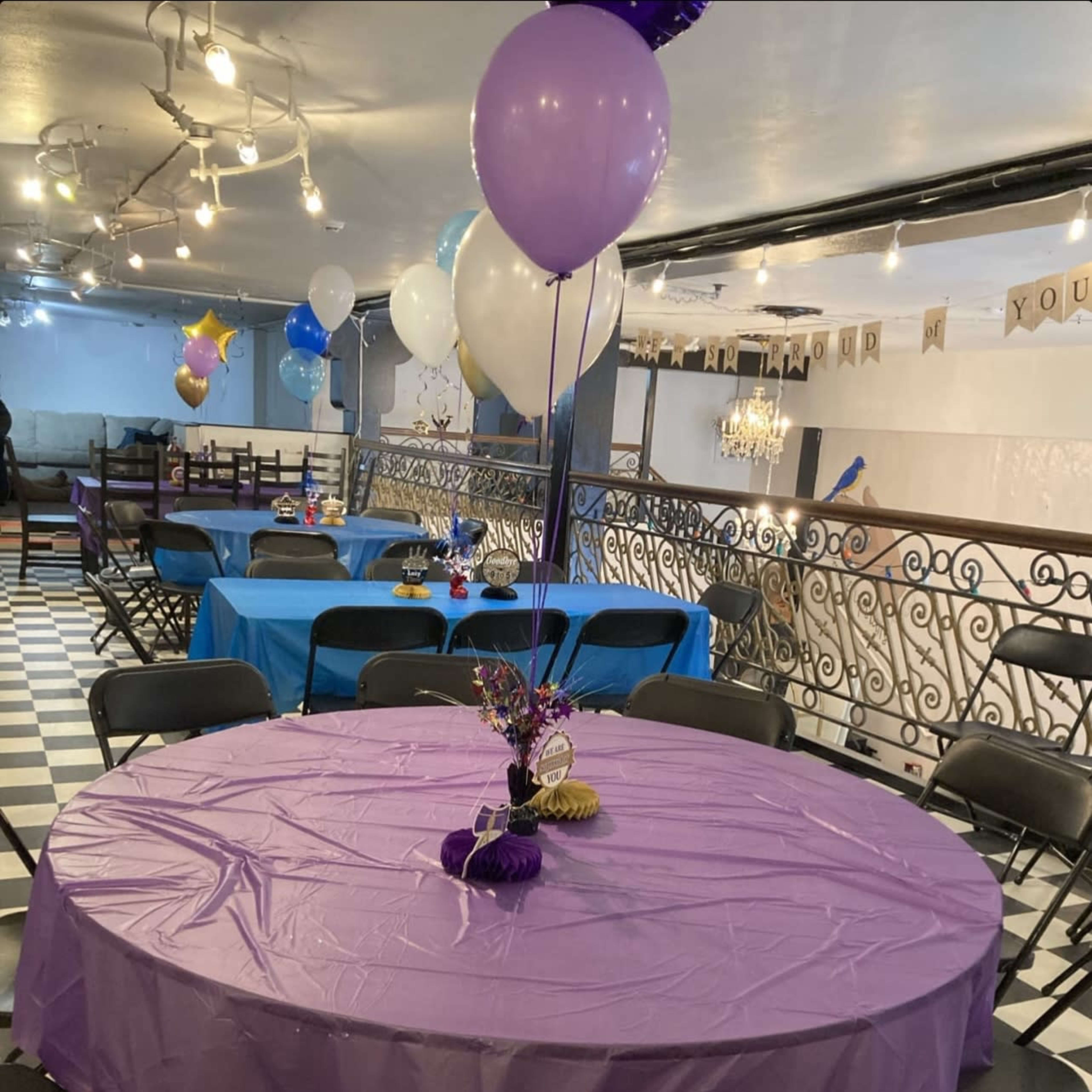 A round table covered with a purple tablecloth is surrounded by black chairs in a room decorated with balloons and streamers.