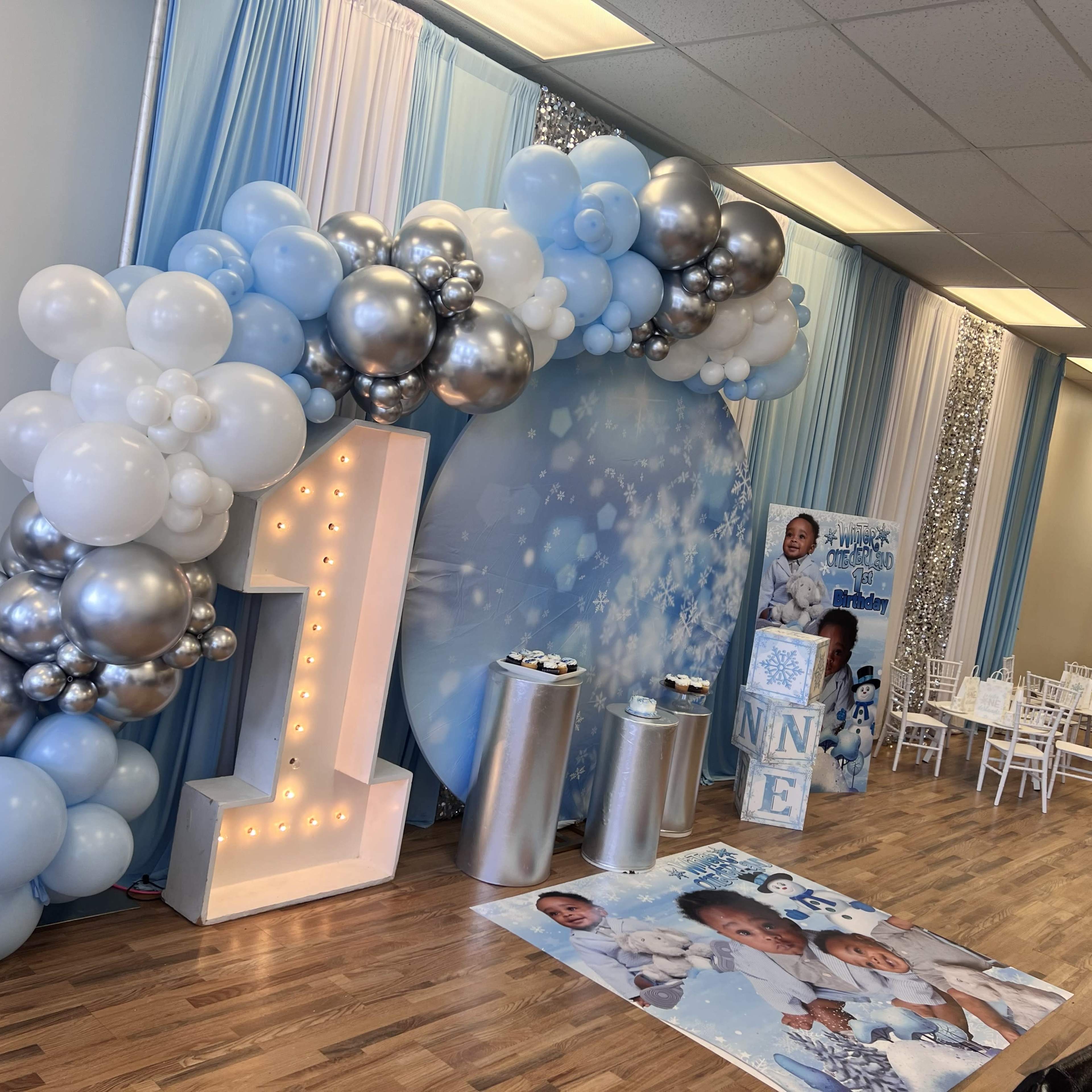 The scene features a birthday party setup with a large illuminated number "1" surrounded by blue and silver balloons, a decorative backdrop, and tables arranged for guests.