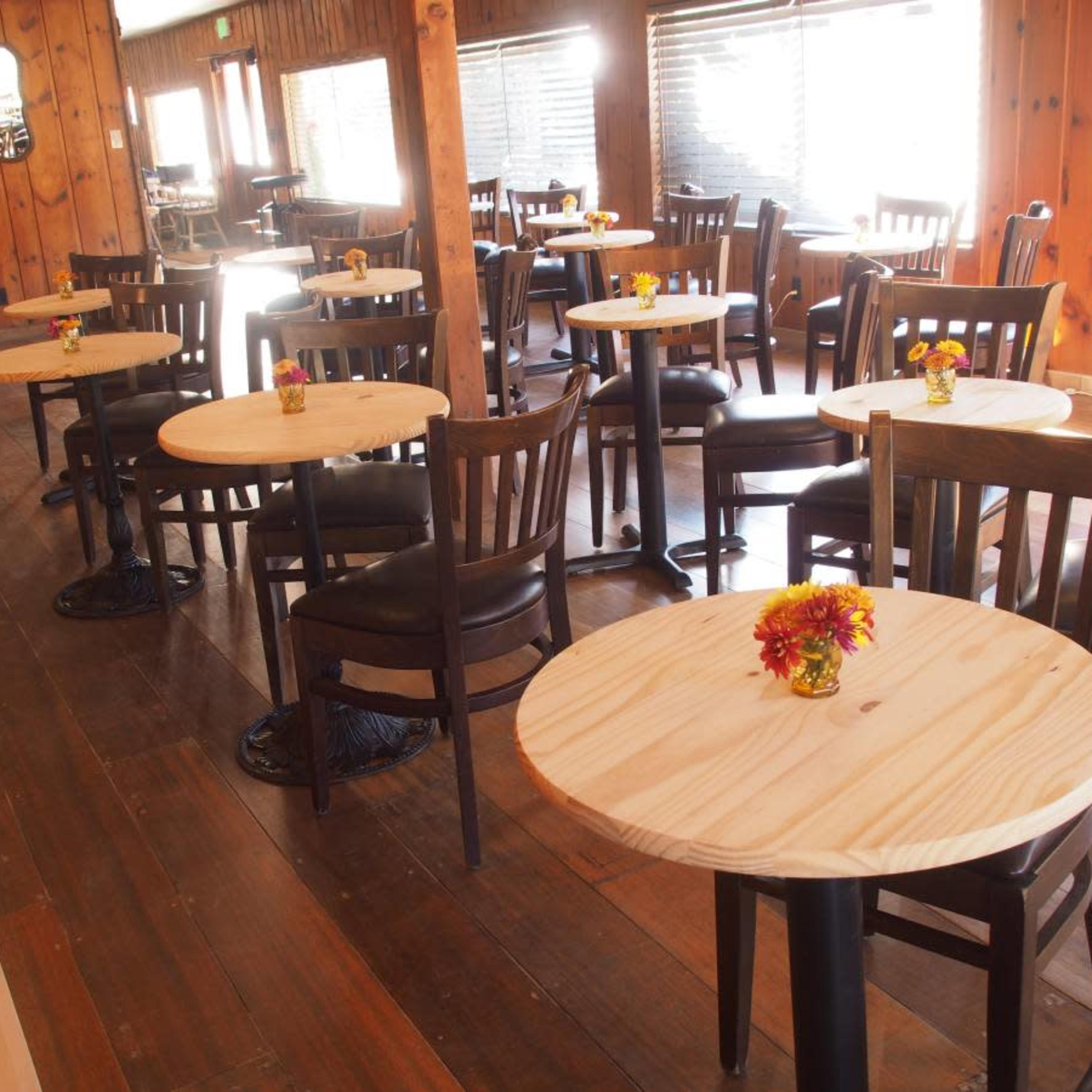A wooden restaurant interior with multiple small round tables set with flower vases and surrounded by wooden chairs.