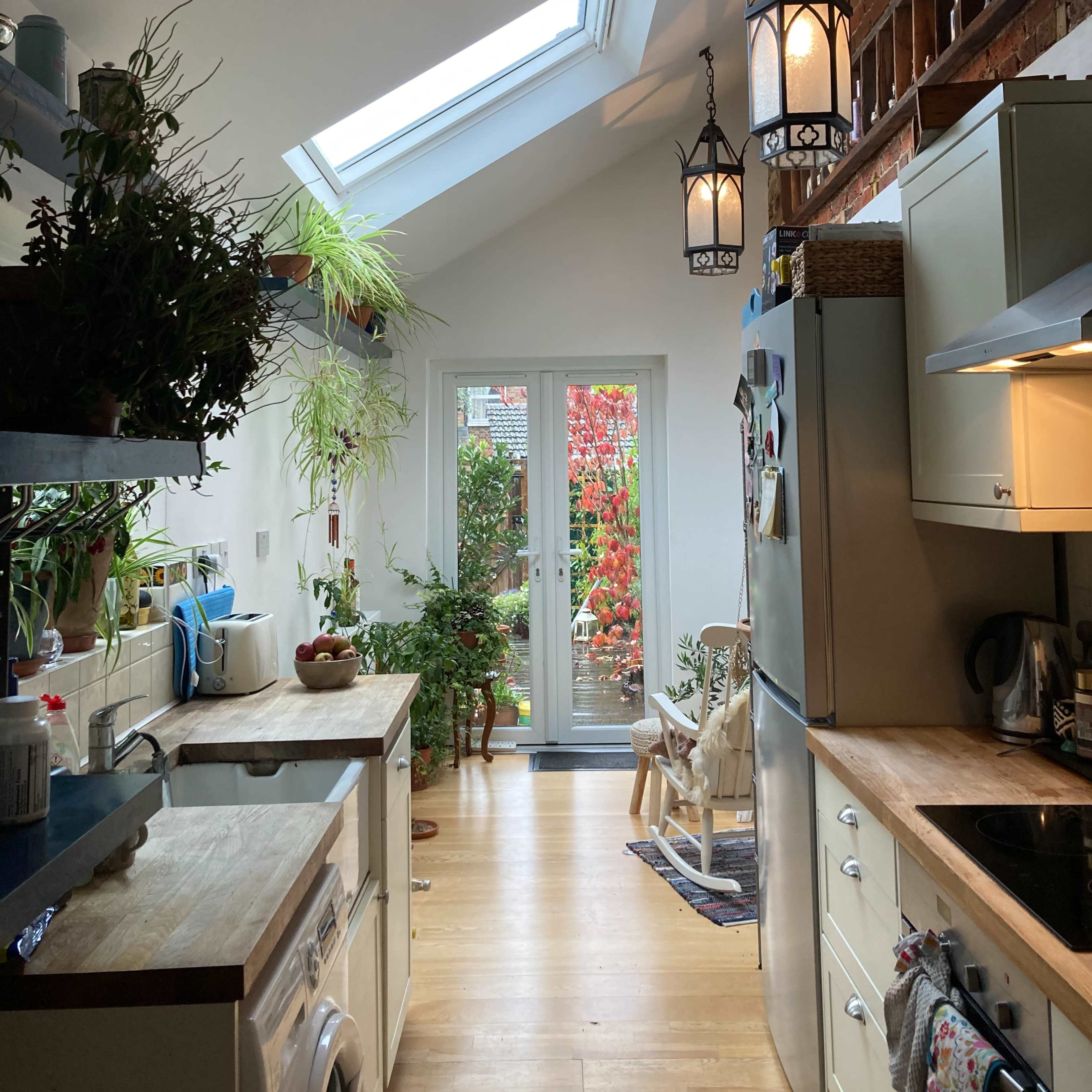 A well-lit kitchen features a skylight, potted plants, and various appliances, leading to a glass door that opens to the garden.