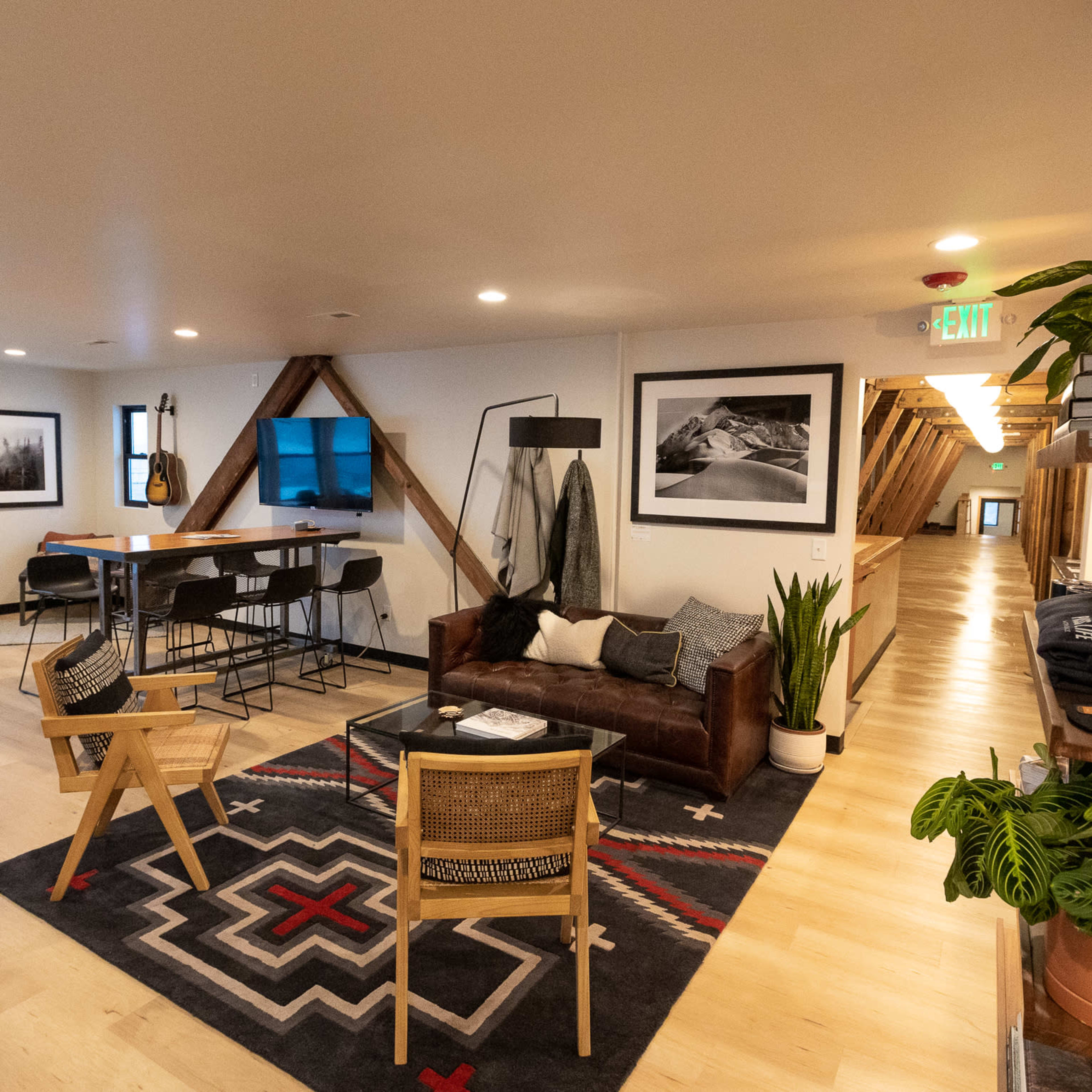 A modern open-concept lounge area featuring a leather sofa, wooden chairs, and a rug, with a dining space and hallway in the background.