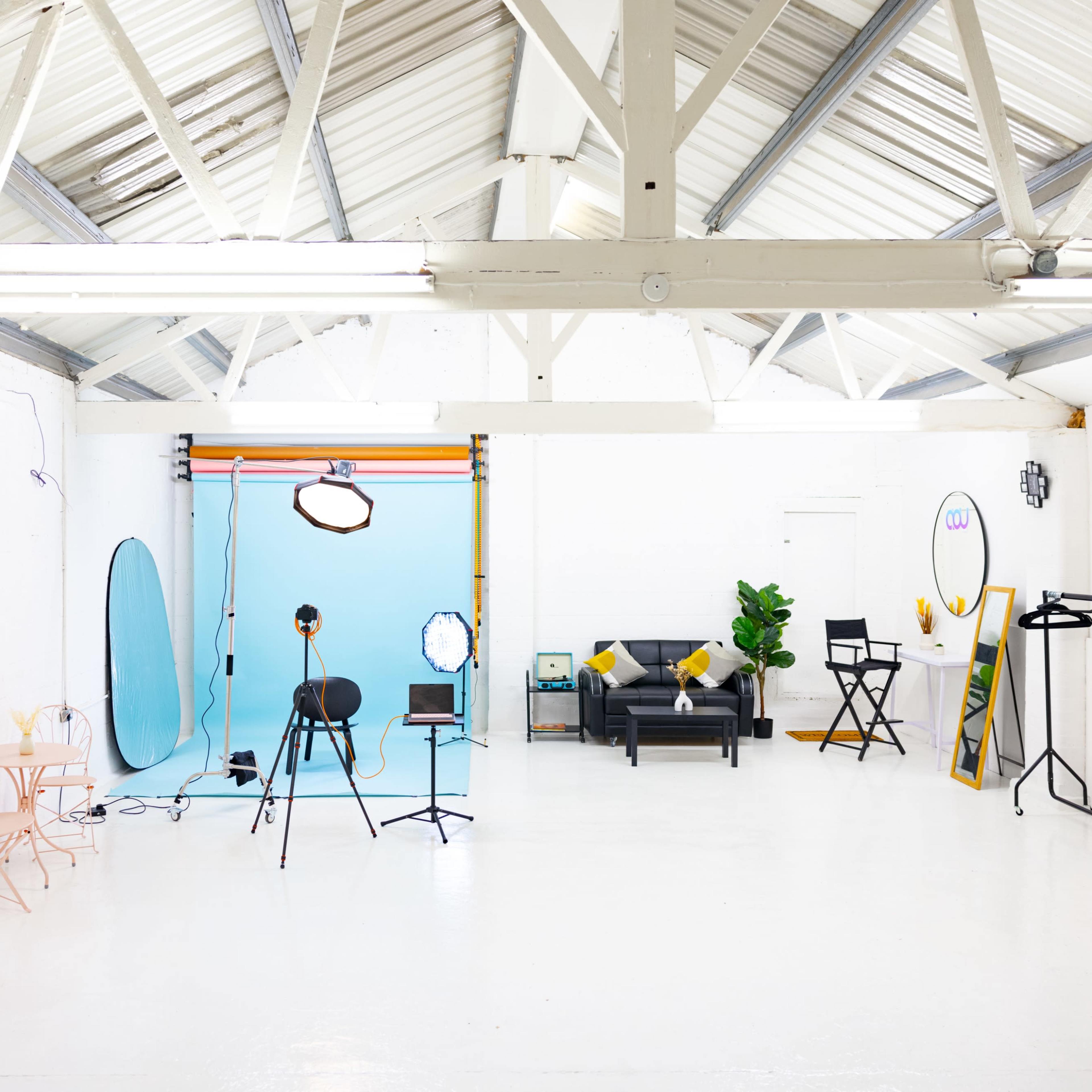 A spacious, well-lit studio features a blue backdrop, various photography equipment, and furniture including a black couch and a pink table set.