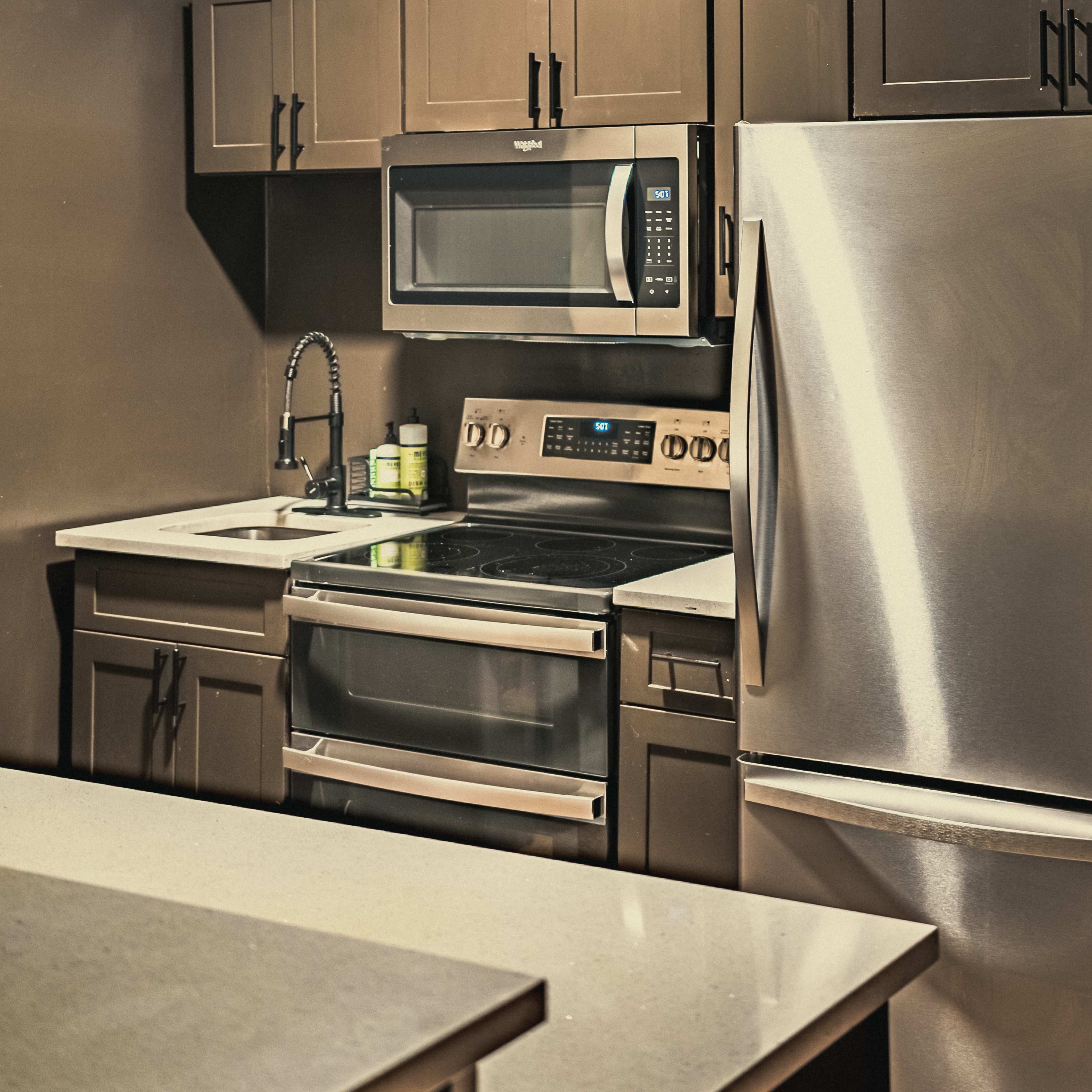A compact kitchen featuring dark cabinets, a stainless steel refrigerator, a microwave above an electric stove, and a sink with a faucet.