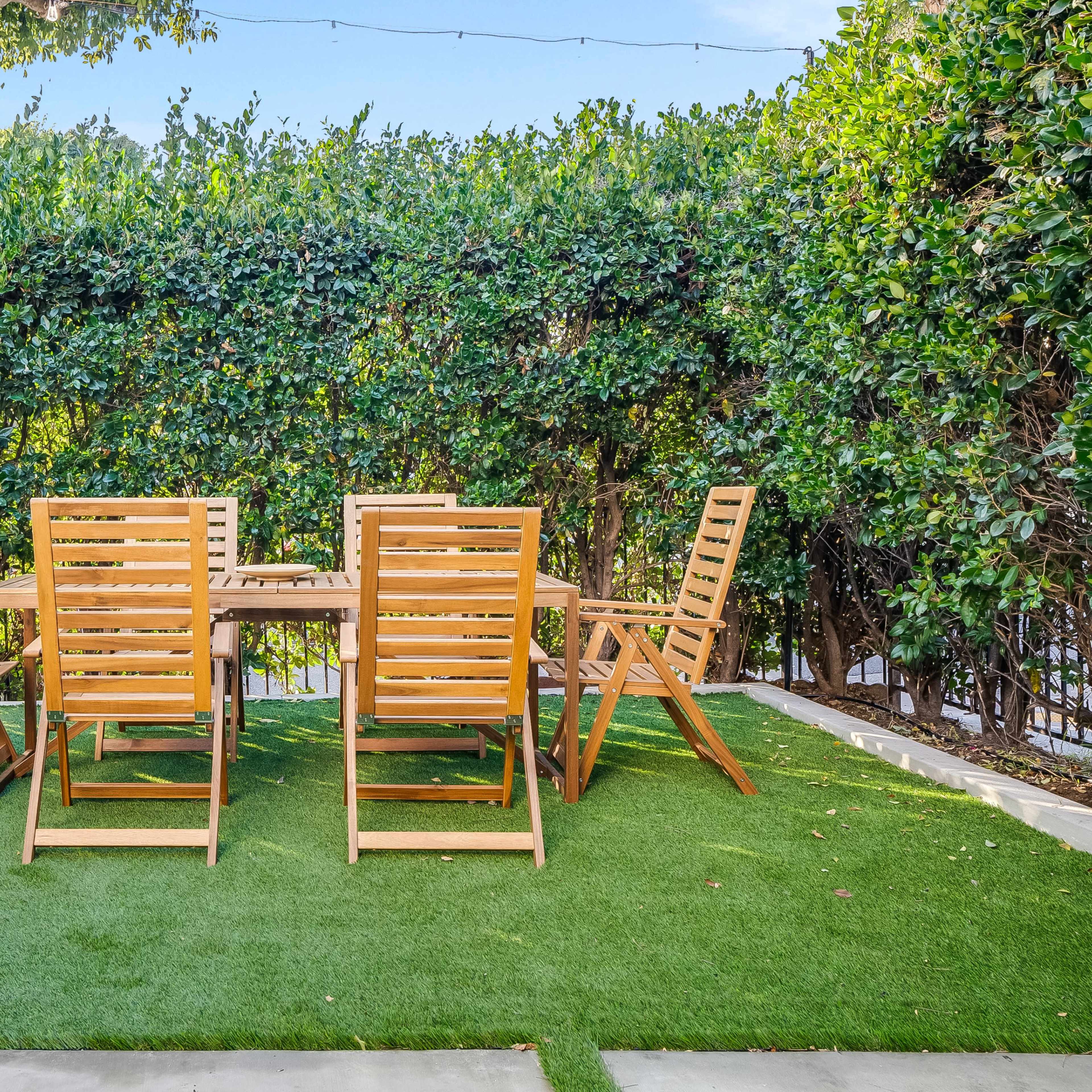 A wooden dining table with six folding chairs is set on a green lawn surrounded by tall hedges.