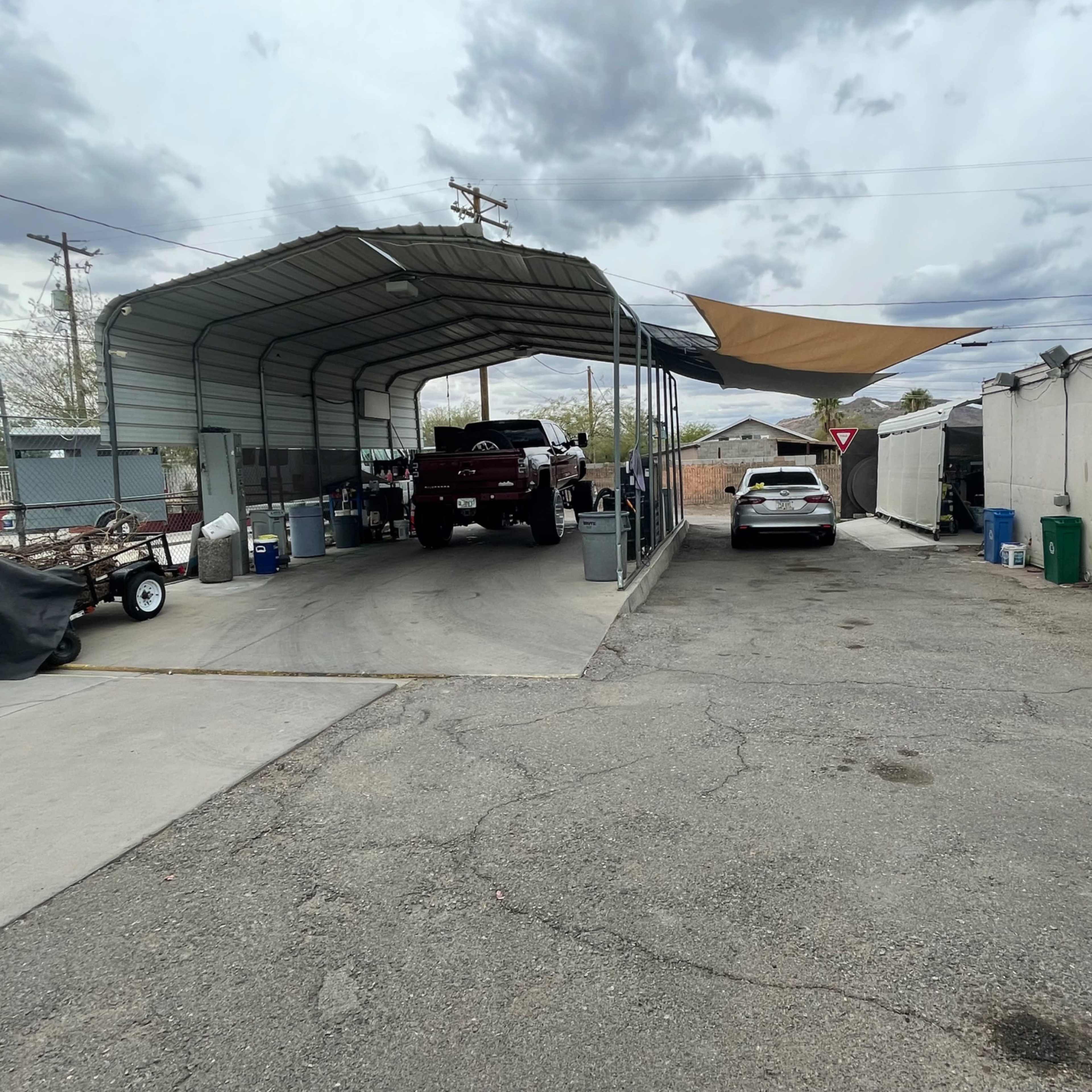 A covered parking area with two vehicles, one under a metal awning and another parked in an open space, alongside a dirt driveway and various outdoor items.