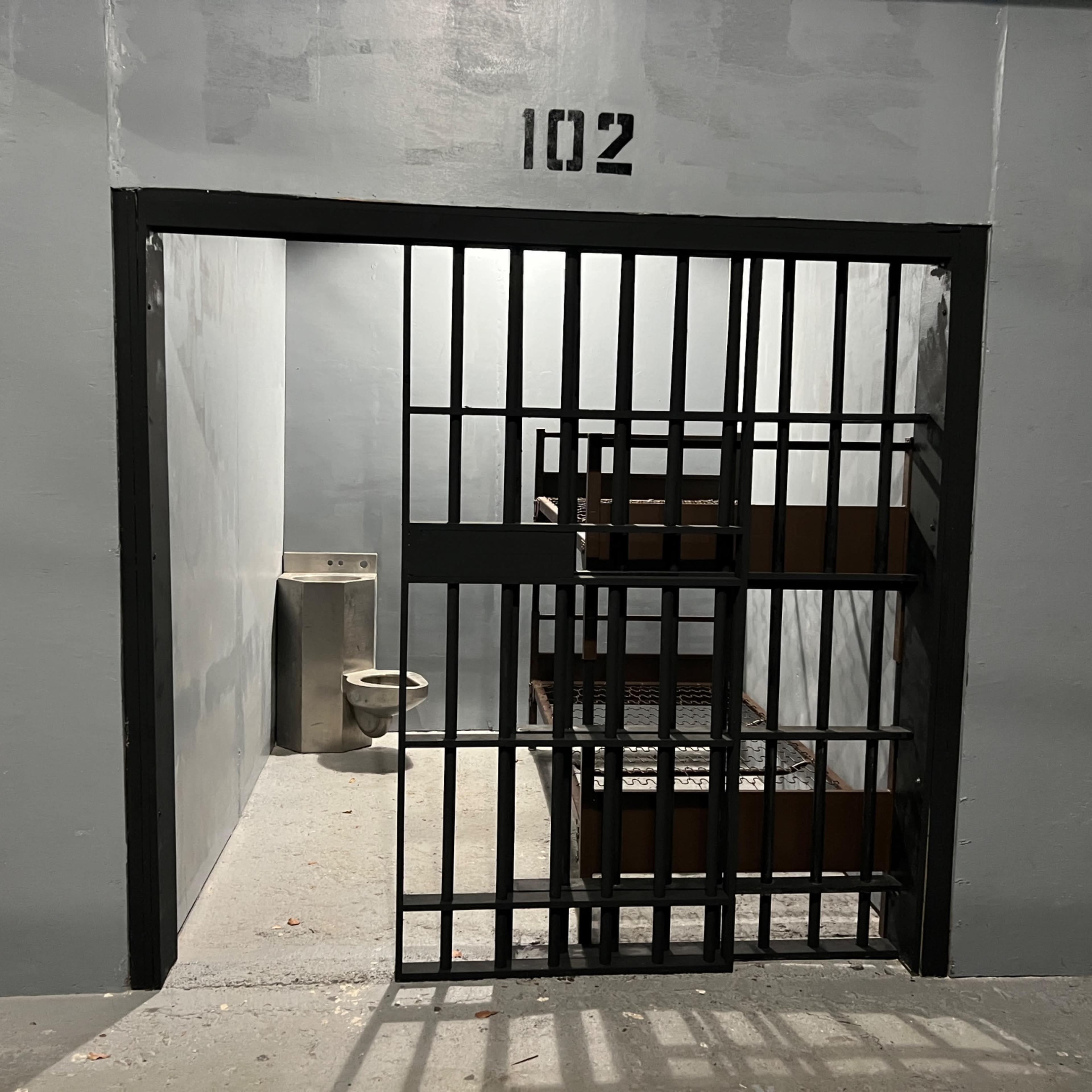An open jail cell marked with the number 102, featuring a toilet and two metal bunks.