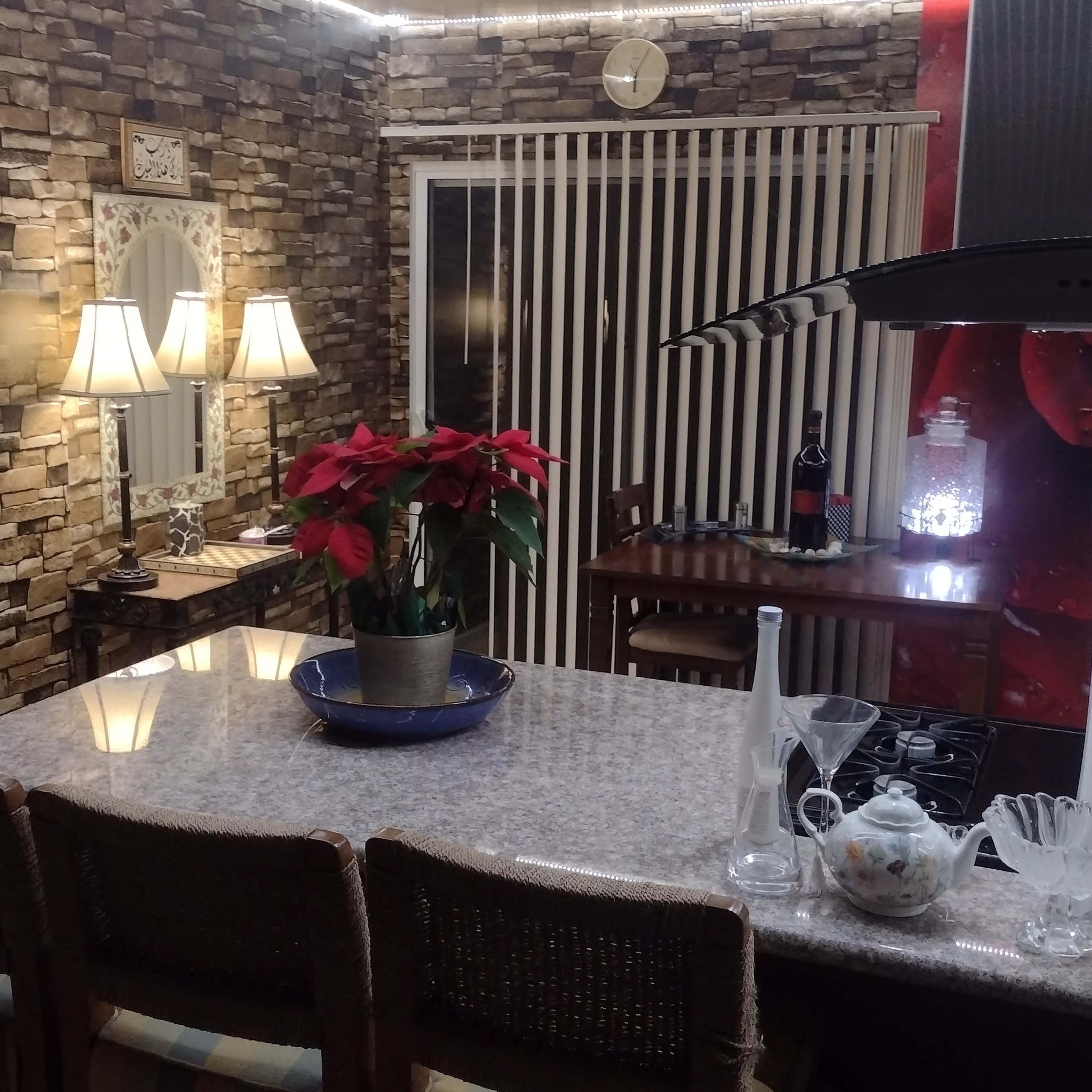 A kitchen with stone-patterned walls, a granite countertop with three stools, and a decorative arrangement of flowers and glassware.