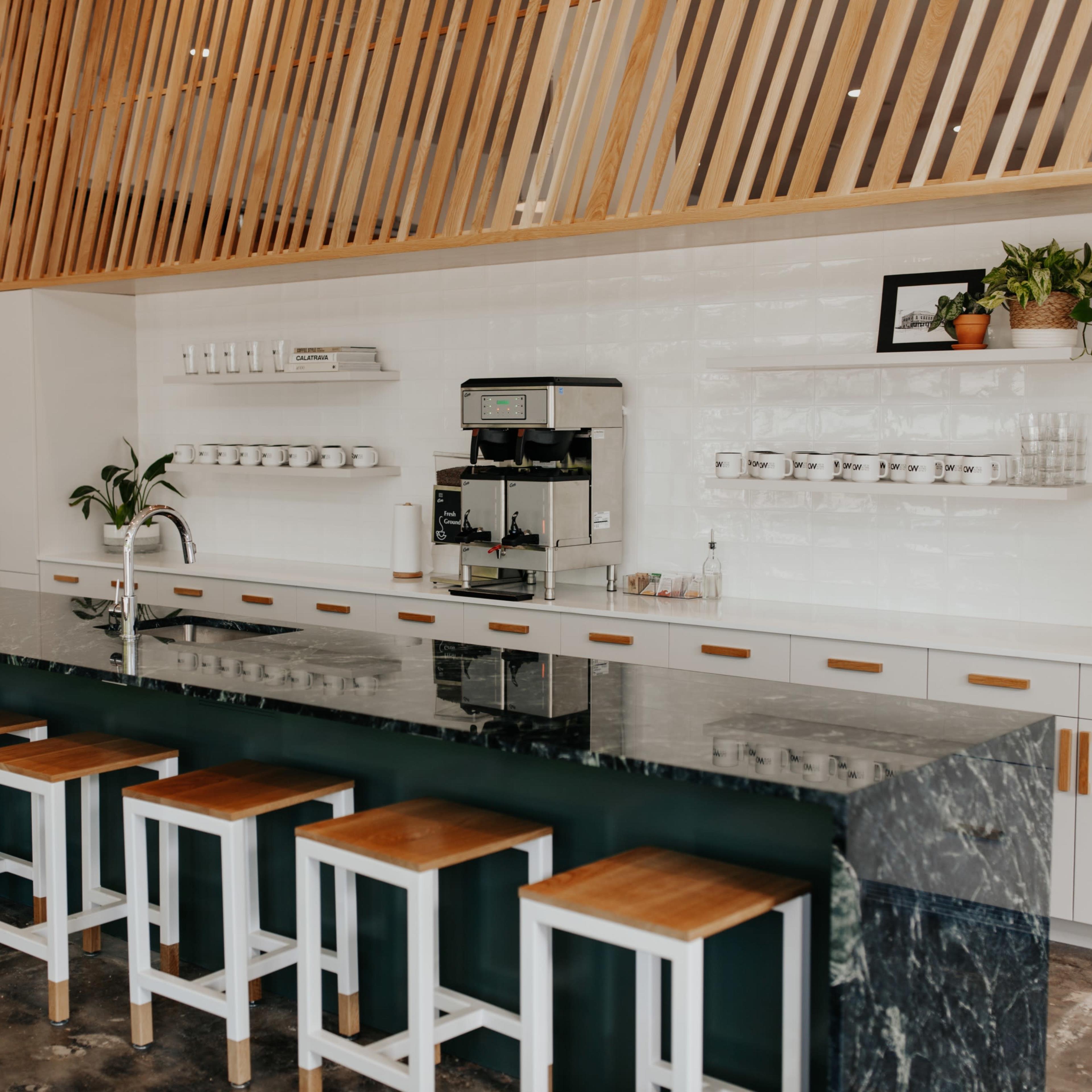 A modern cafe interior features a long marble counter with wooden stools, a coffee machine, and neatly arranged shelves above.