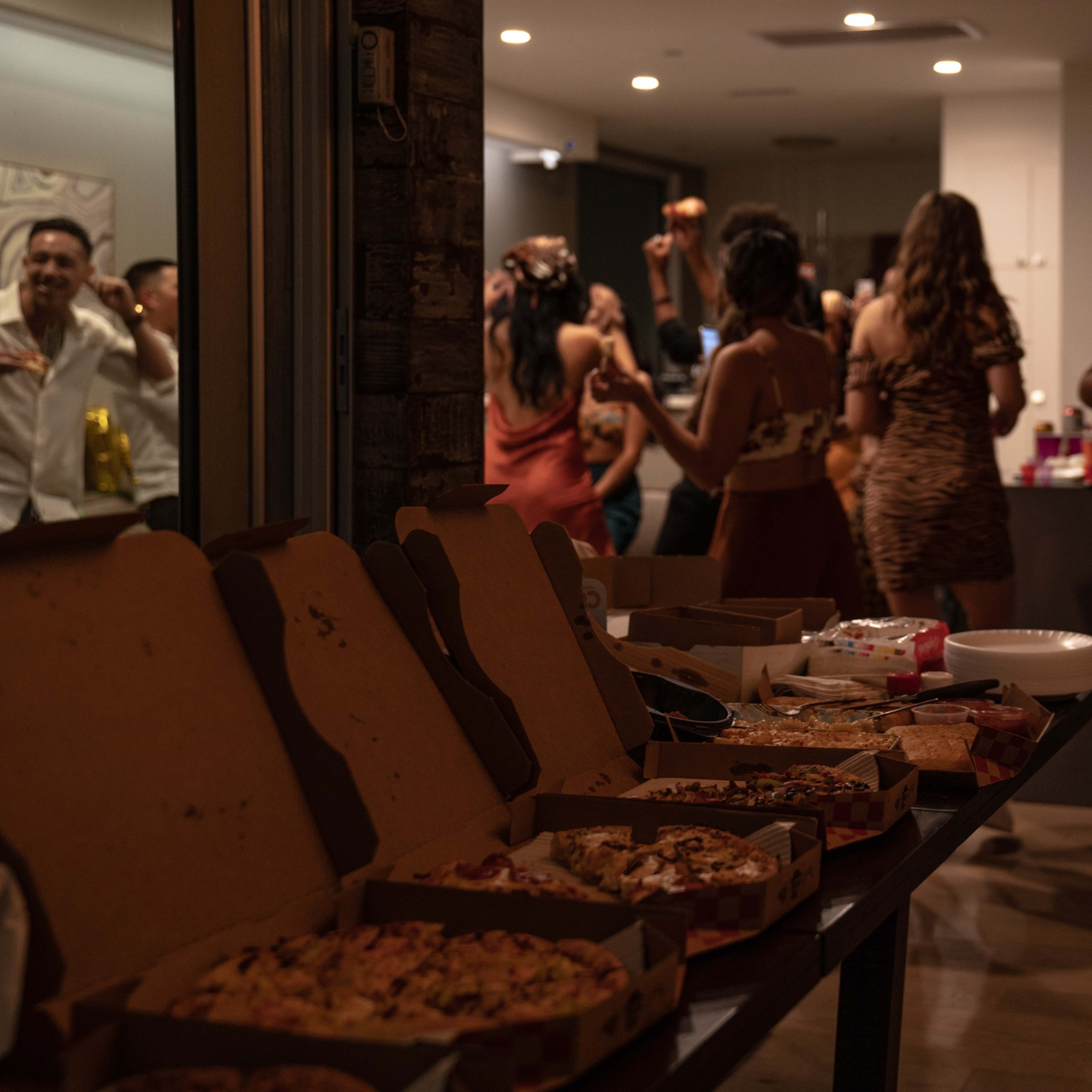 A gathering of people is enjoying a party in a modern space, with several pizza boxes and food laid out in the foreground.