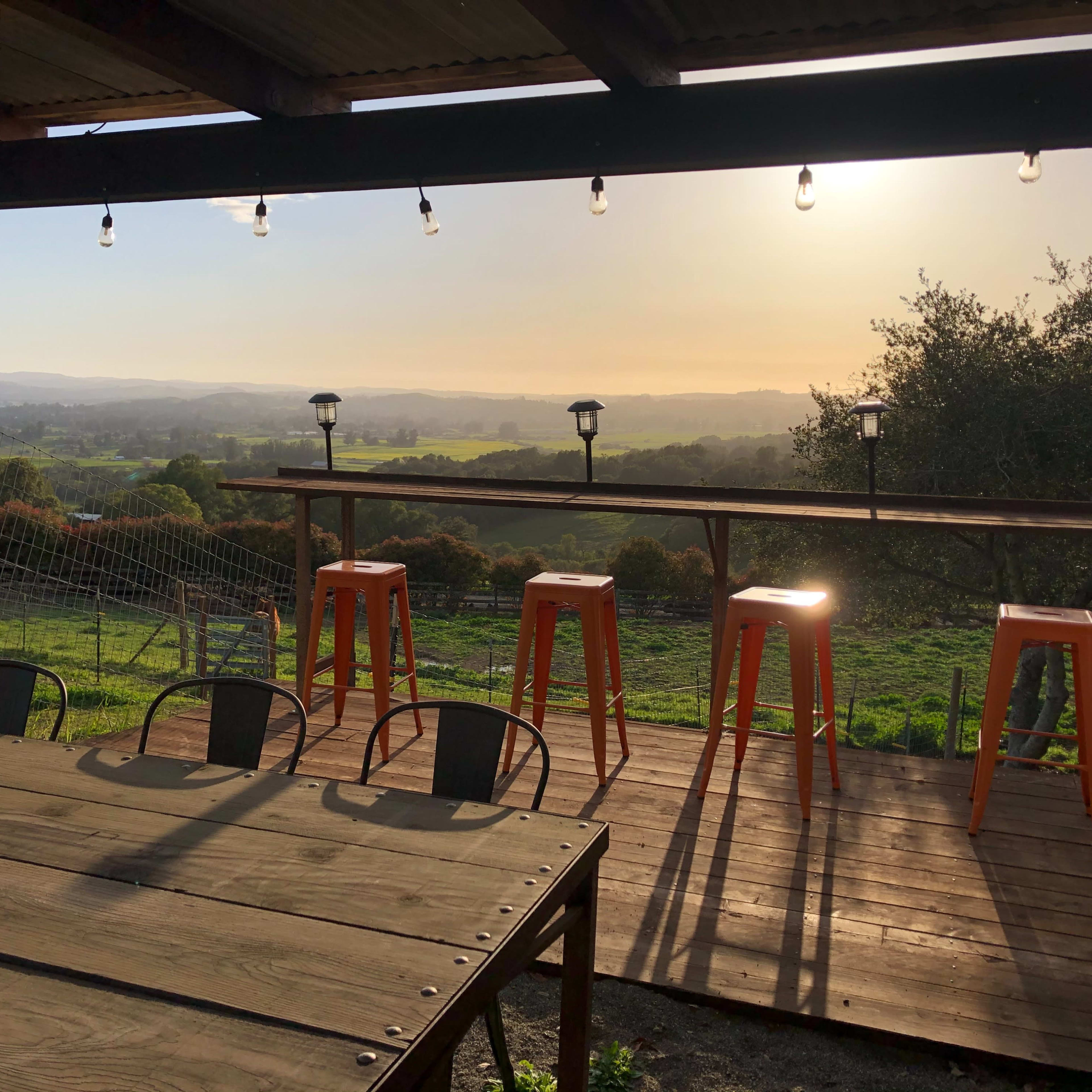 A wooden deck features tall orange stools with a scenic view of rolling hills at sunset.