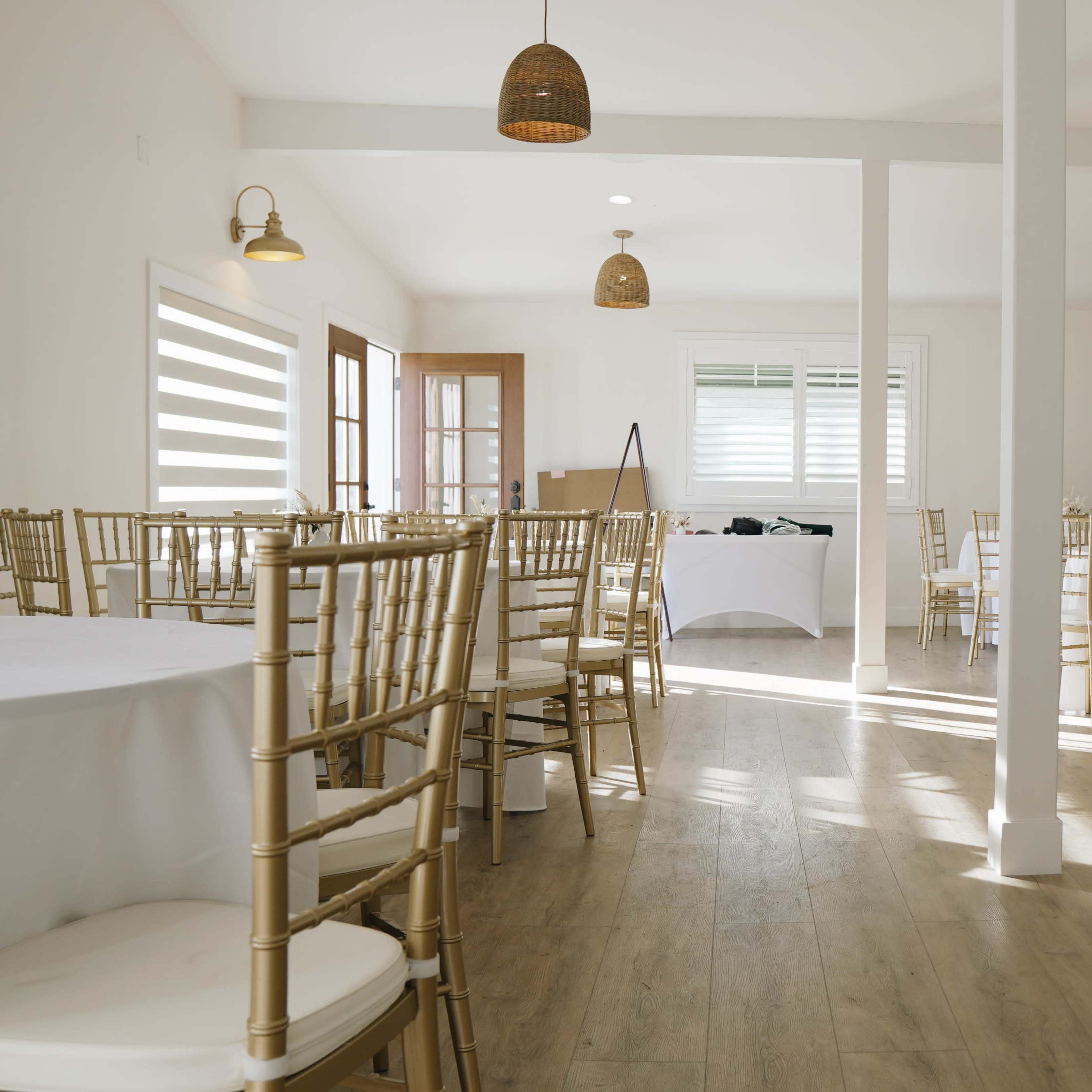 A spacious, brightly lit event room with wooden floors and tables set with white linens and gold chairs, complemented by natural light from large windows.