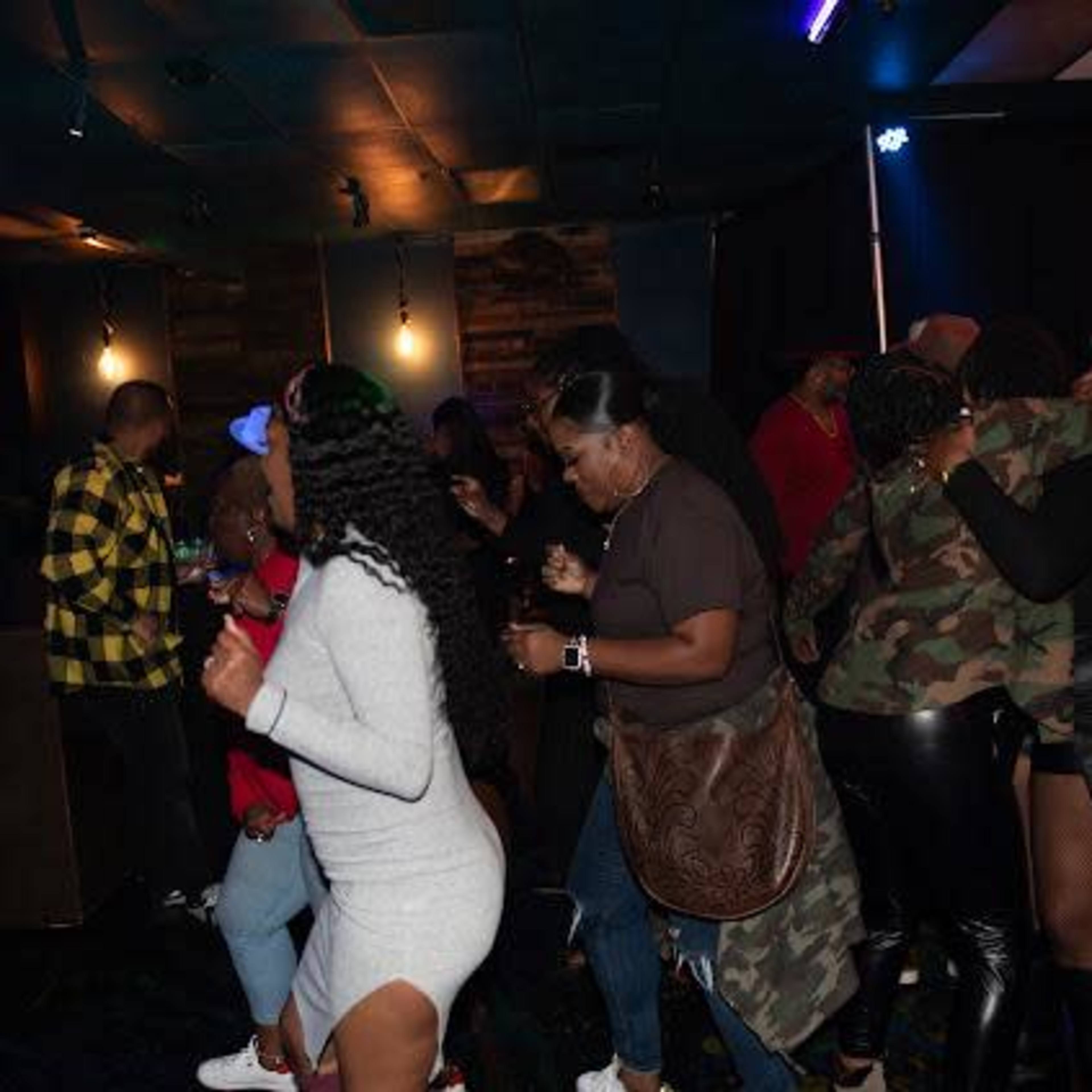 A group of people dance in a dimly lit venue, with some wearing diverse outfits while enjoying the lively atmosphere.