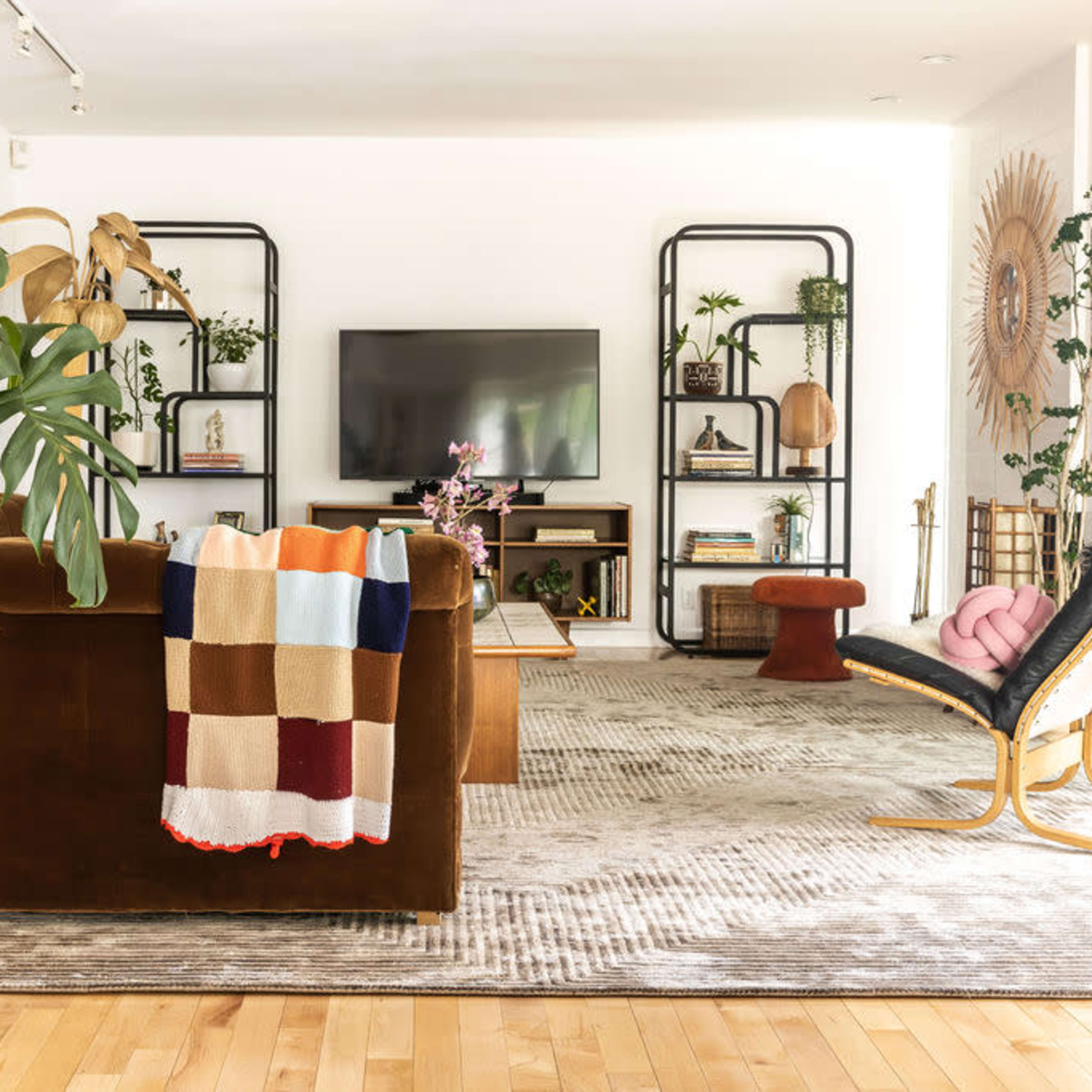 A modern living room with a brown upholstered sofa, a large television mounted on the wall, and various decorative shelves filled with plants and accessories.