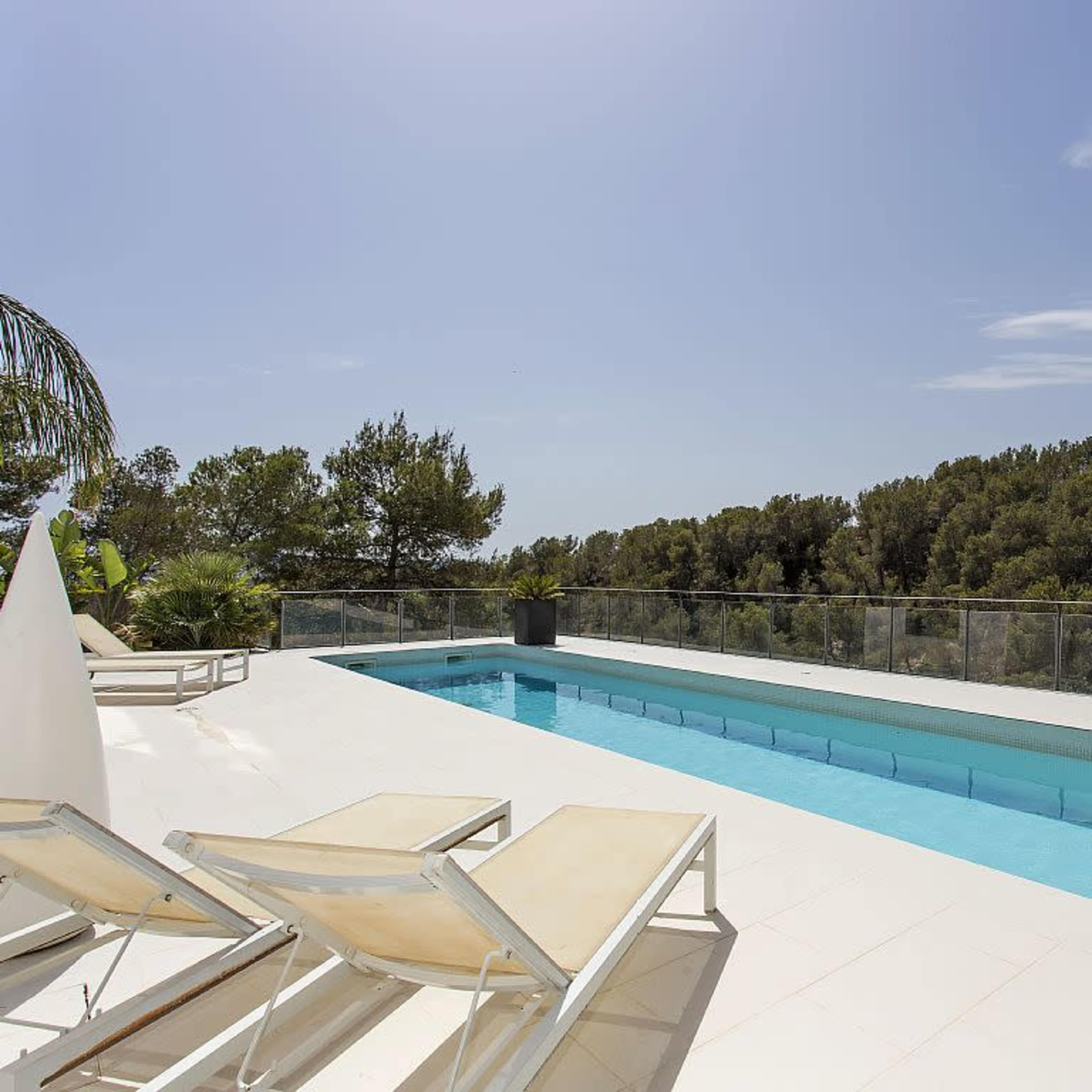 A clean, modern pool area features lounge chairs beside a rectangular swimming pool, surrounded by greenery and clear blue skies.
