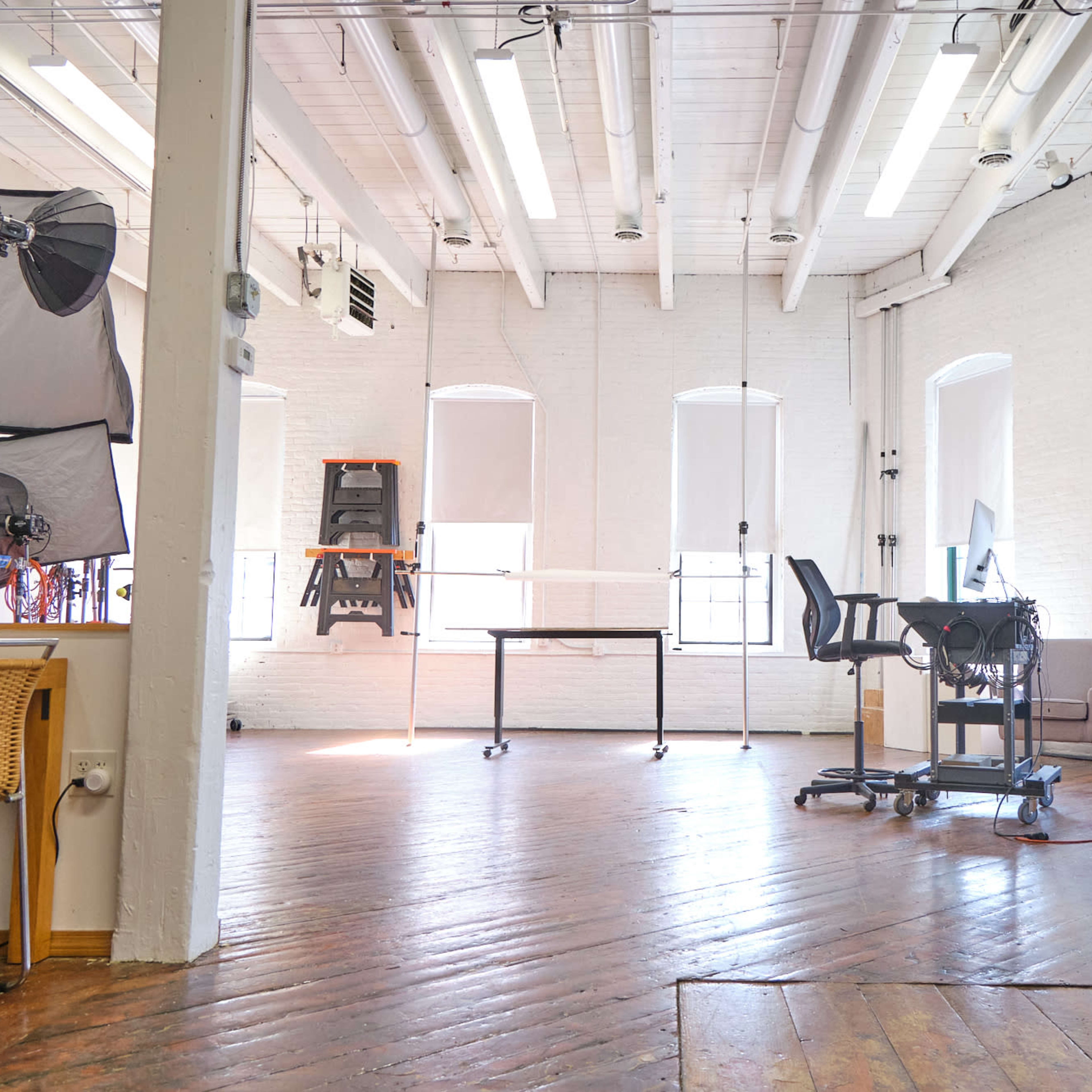 A bright, open workspace featuring wooden floors, large windows with shades, and various furniture including a desk and chairs.