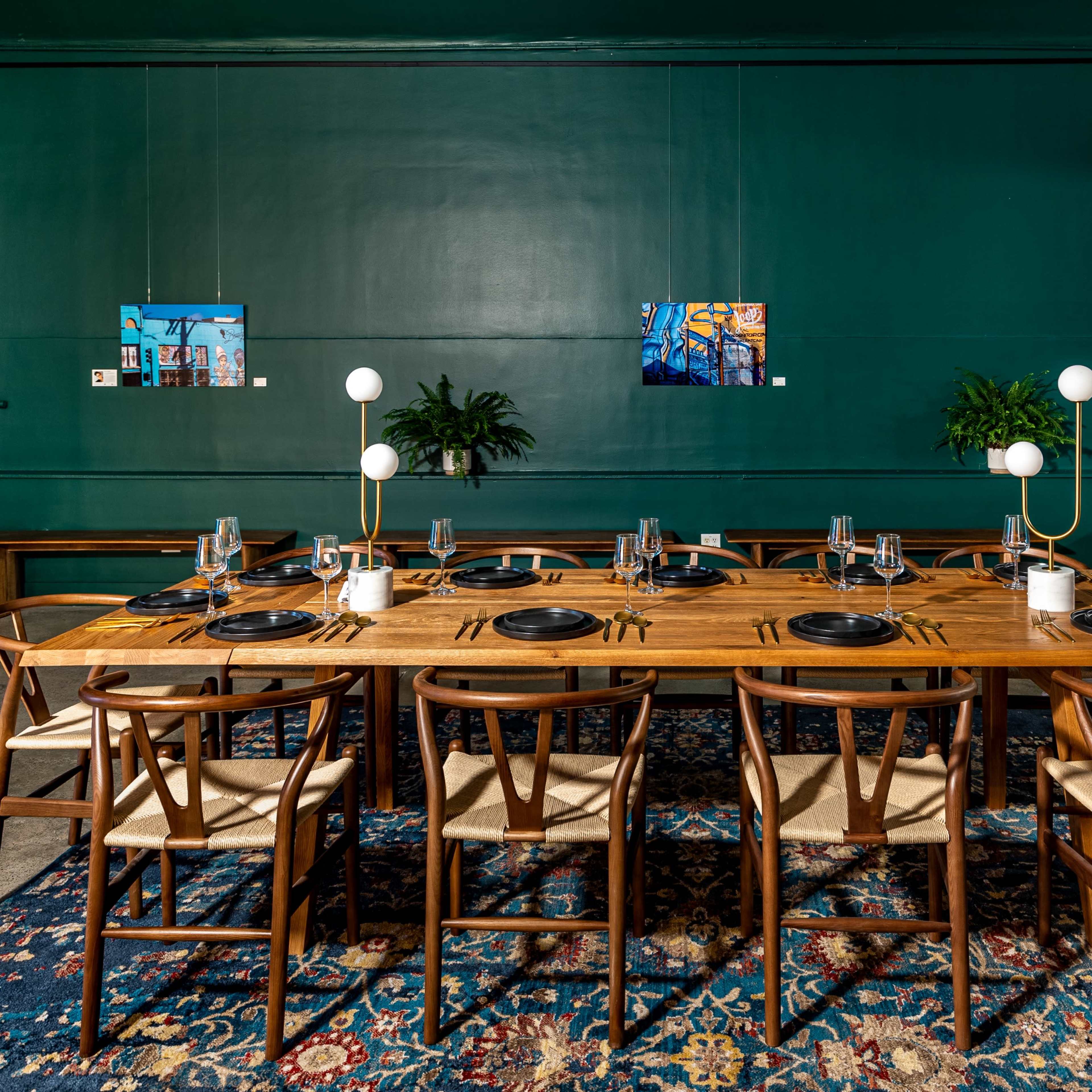 A large wooden dining table is set with plates and glasses, surrounded by chairs, in a room with dark green walls and artwork displayed.