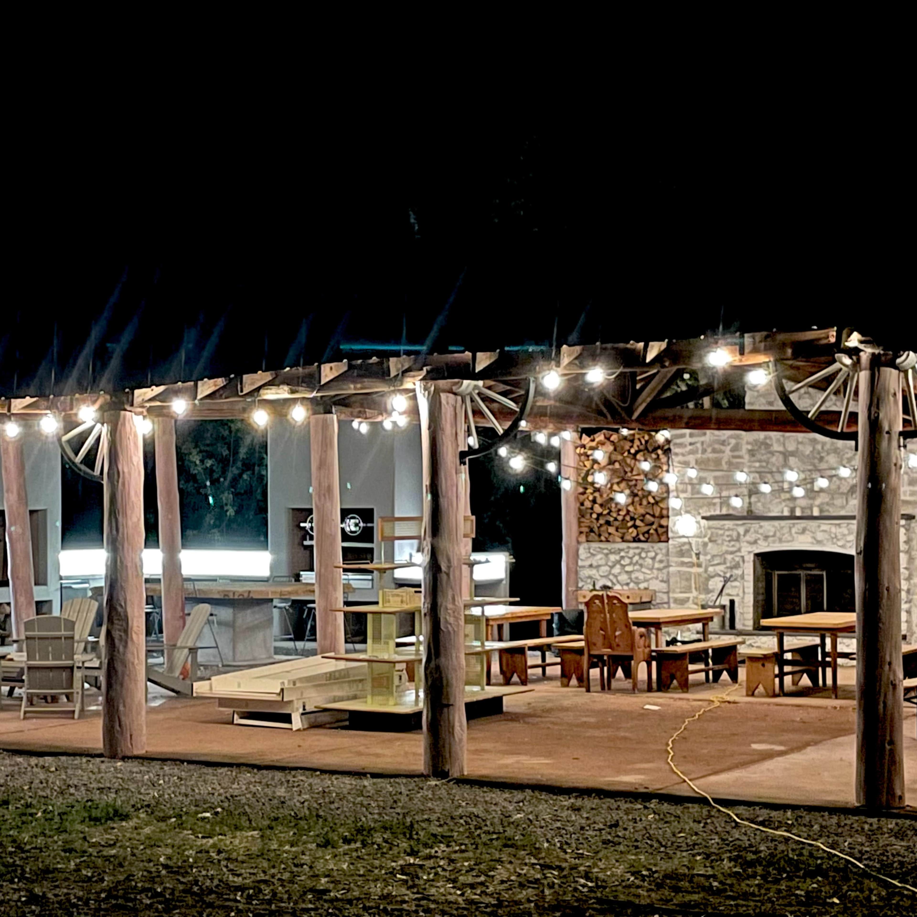 A well-lit outdoor patio features a variety of seating options and a stone fireplace under a wooden pergola at night.