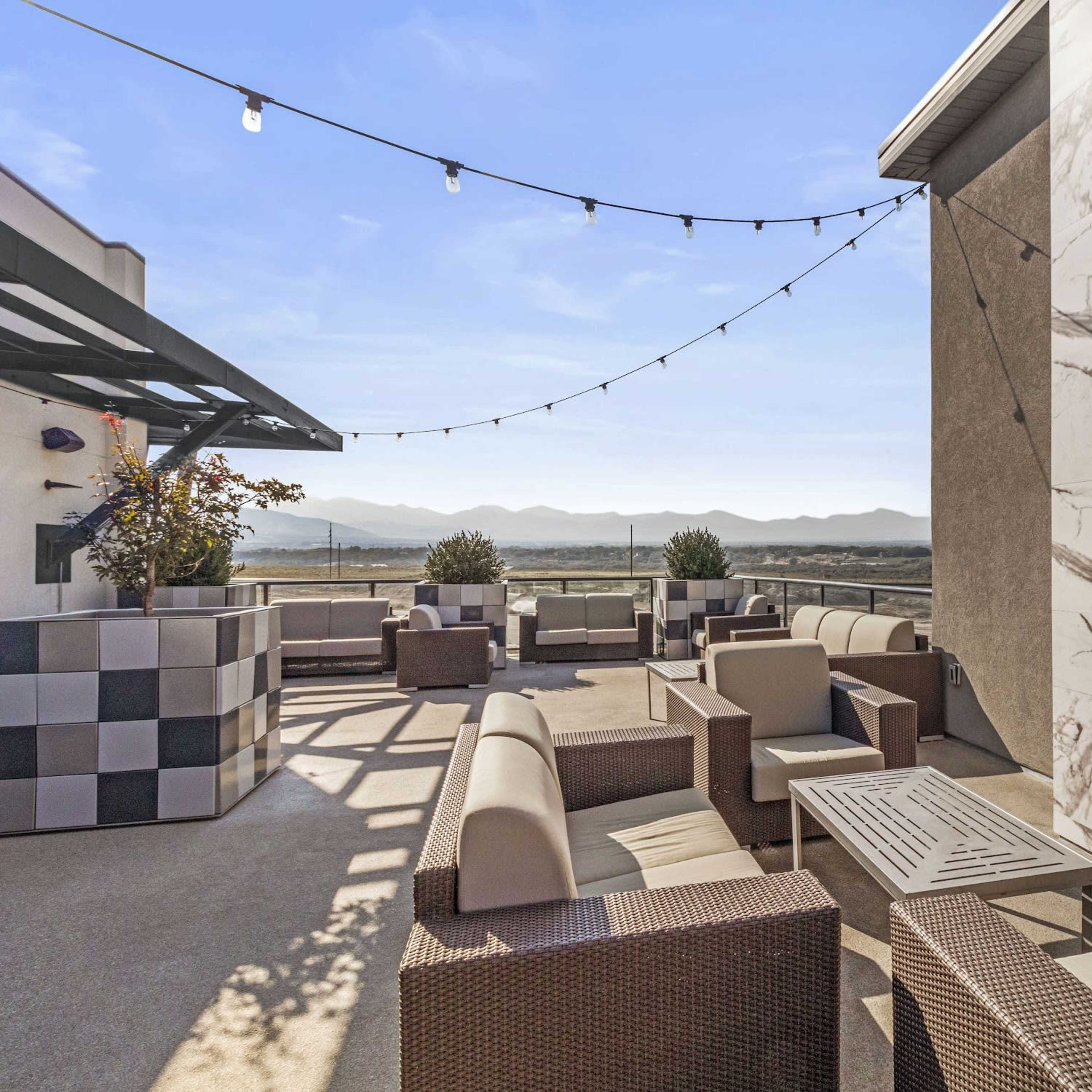 The rooftop terrace features modern seating arrangements, decorative planters, and string lights against a backdrop of mountains.