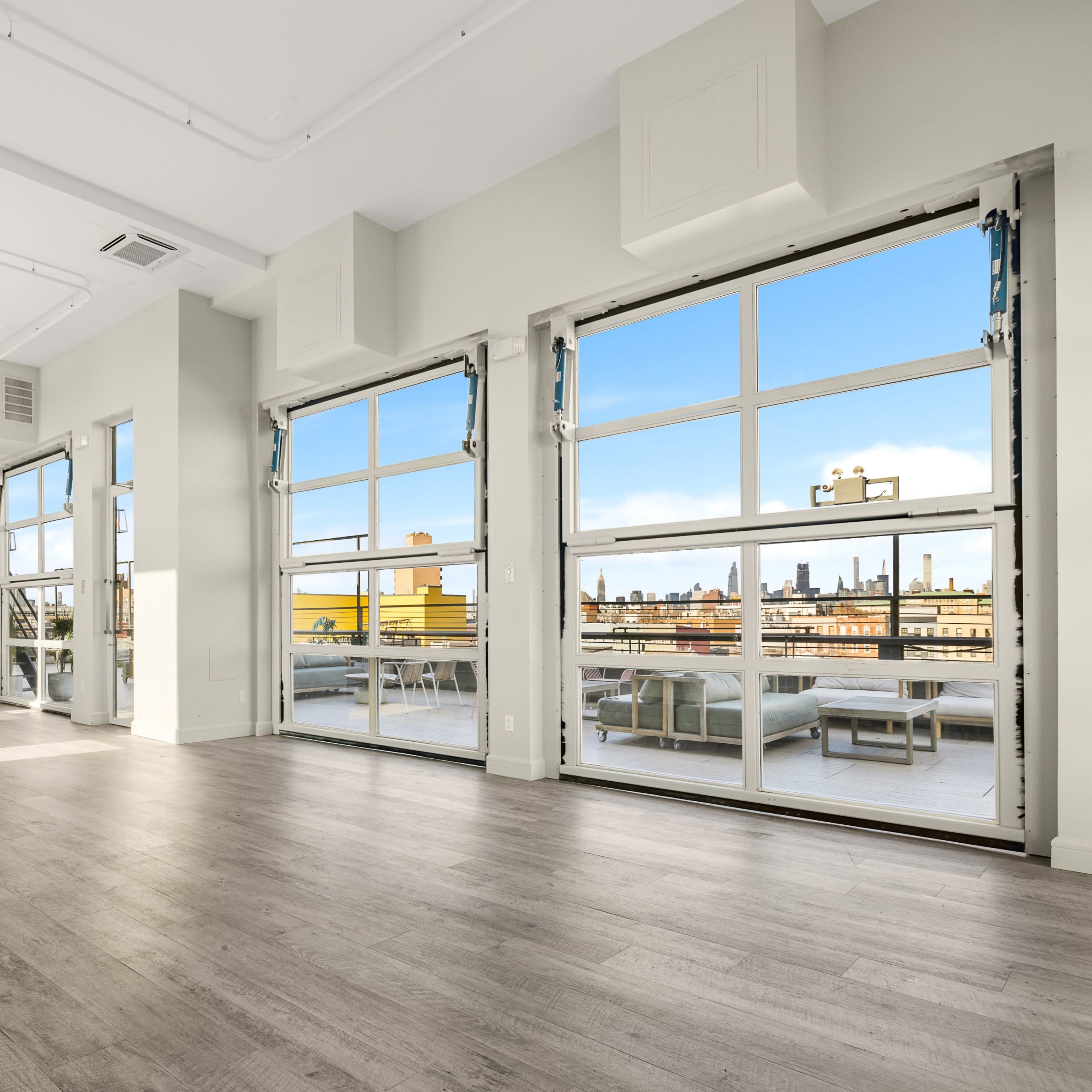 A spacious, modern interior with large windows offering a view of a city skyline and light-colored wooden flooring.