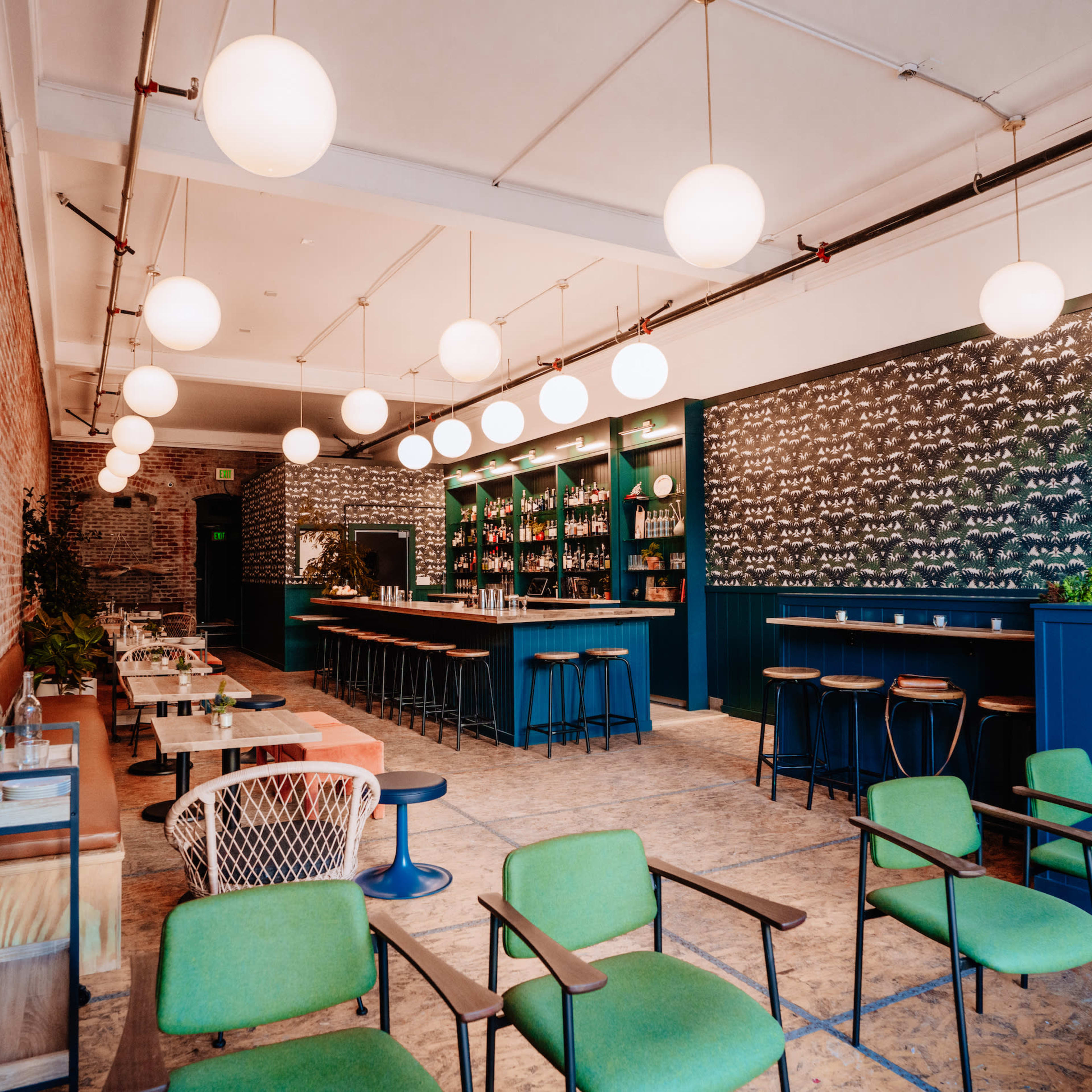 A modern bar interior features green chairs, round pendant lights, and a patterned wall with shelves stocked with bottles.
