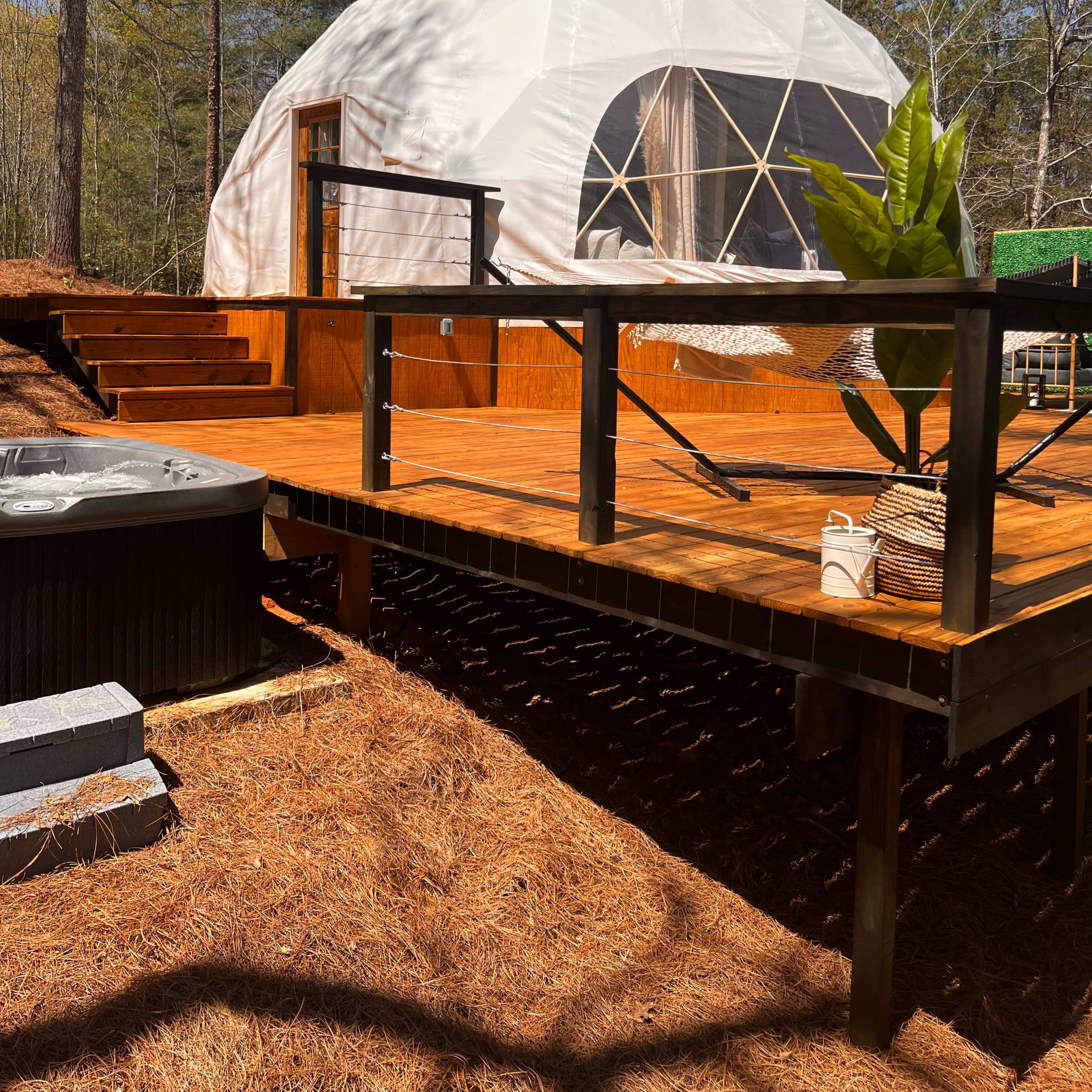 A geodesic dome is situated on a wooden deck surrounded by trees, with a hot tub nearby.