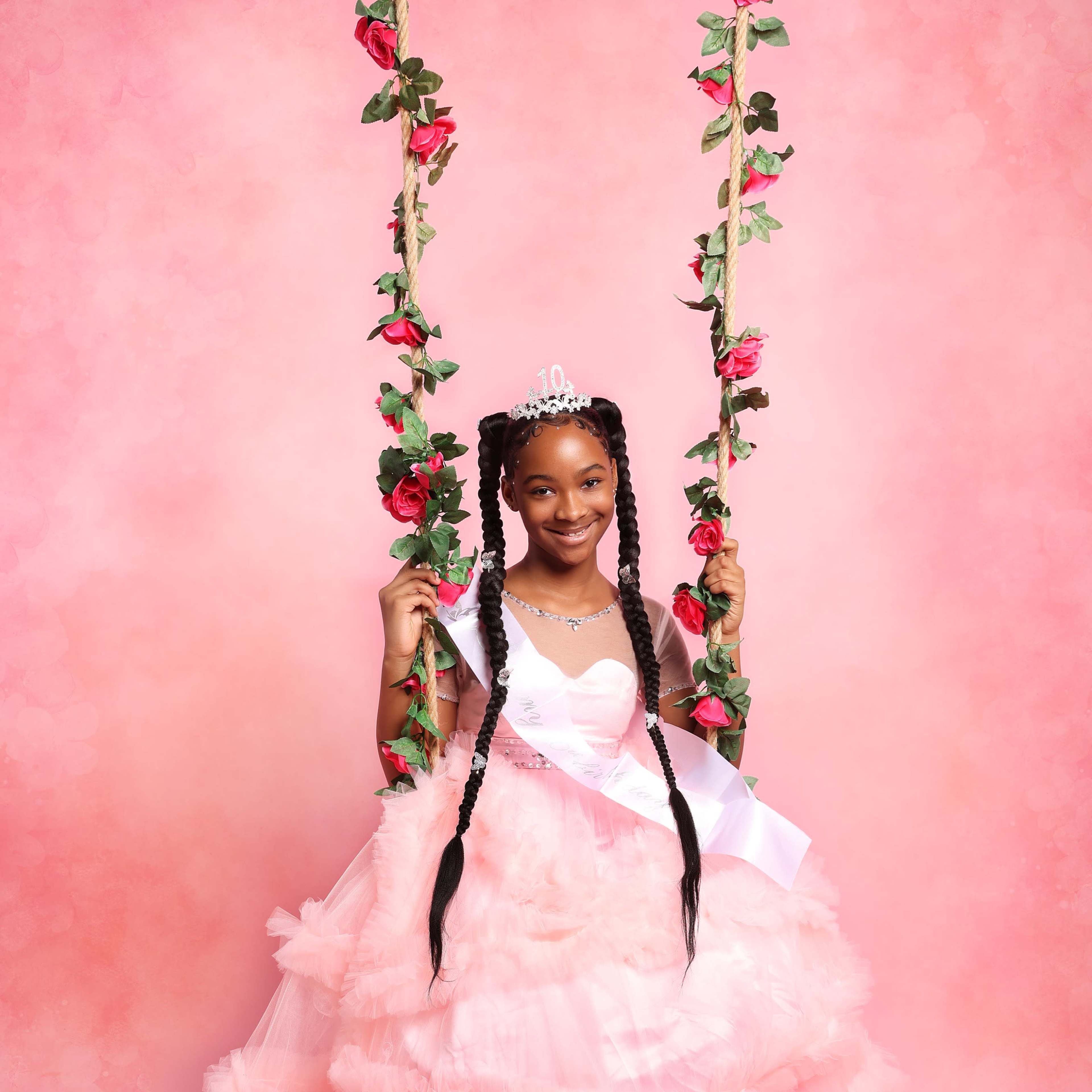 A girl wearing a pink dress with ruffles is standing on a swing decorated with flowers against a pink background.
