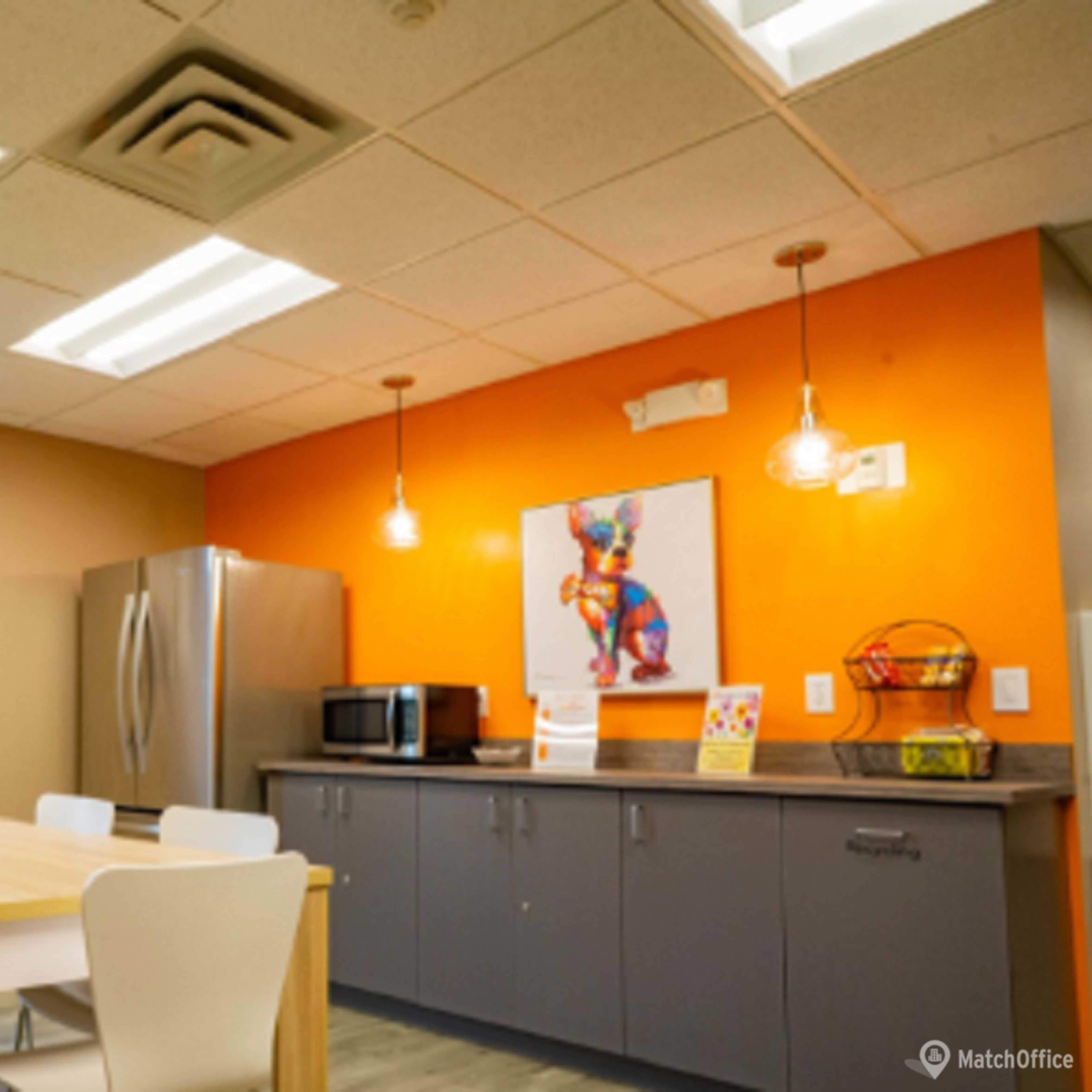 A modern kitchenette with a large table, a refrigerator, a microwave, and colorful artwork on the walls.