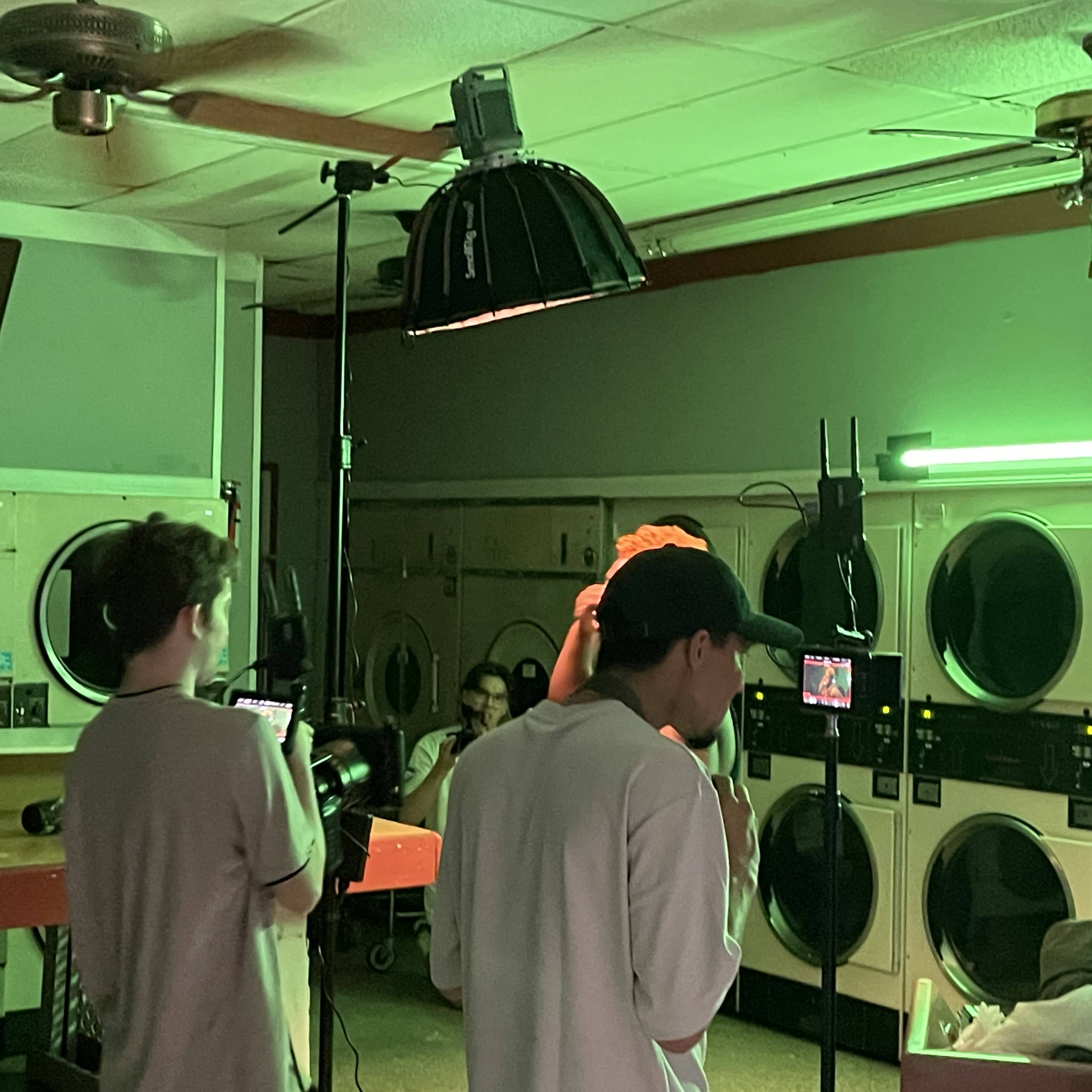 A group of individuals is filming a scene in a laundromat, with washing machines and fluorescent lights in the background.