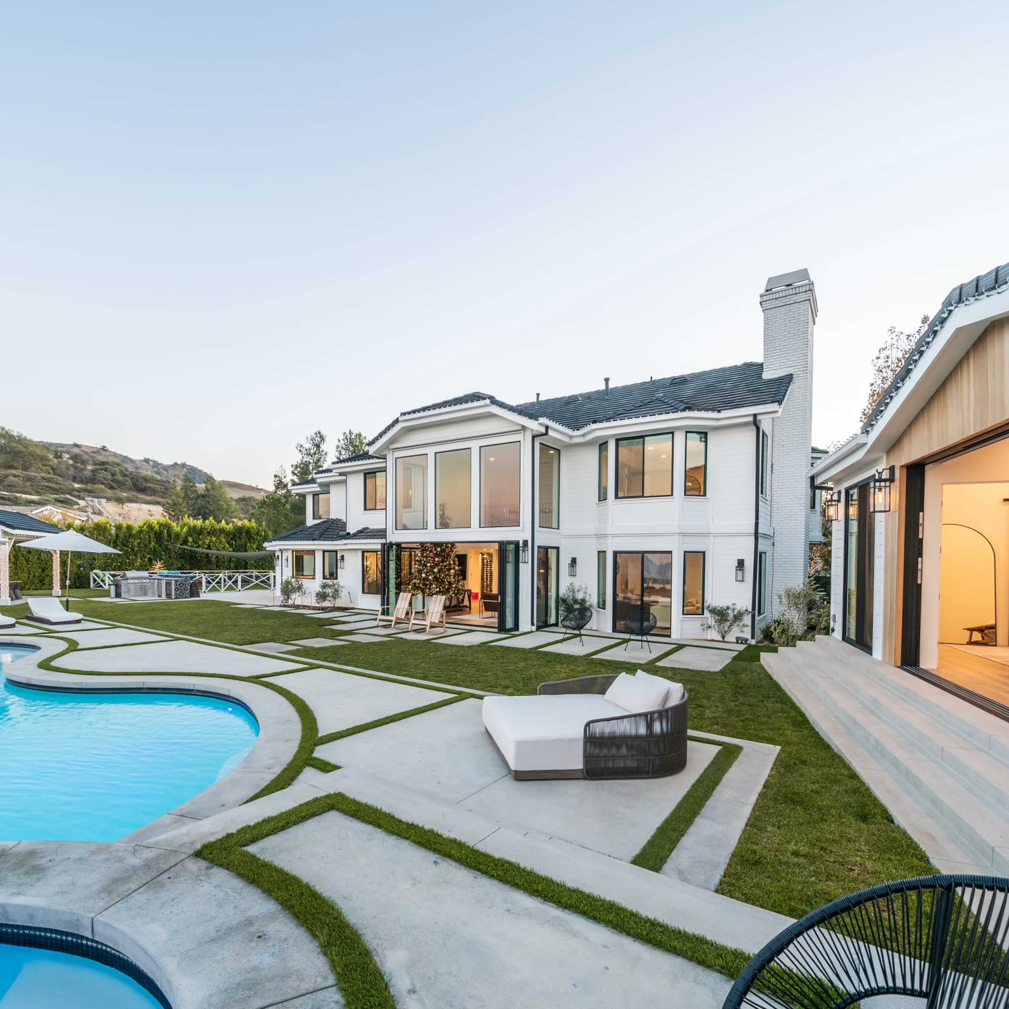 A modern two-story house with large windows, a swimming pool, and a landscaped yard featuring lounge chairs and fitness equipment in a garage.