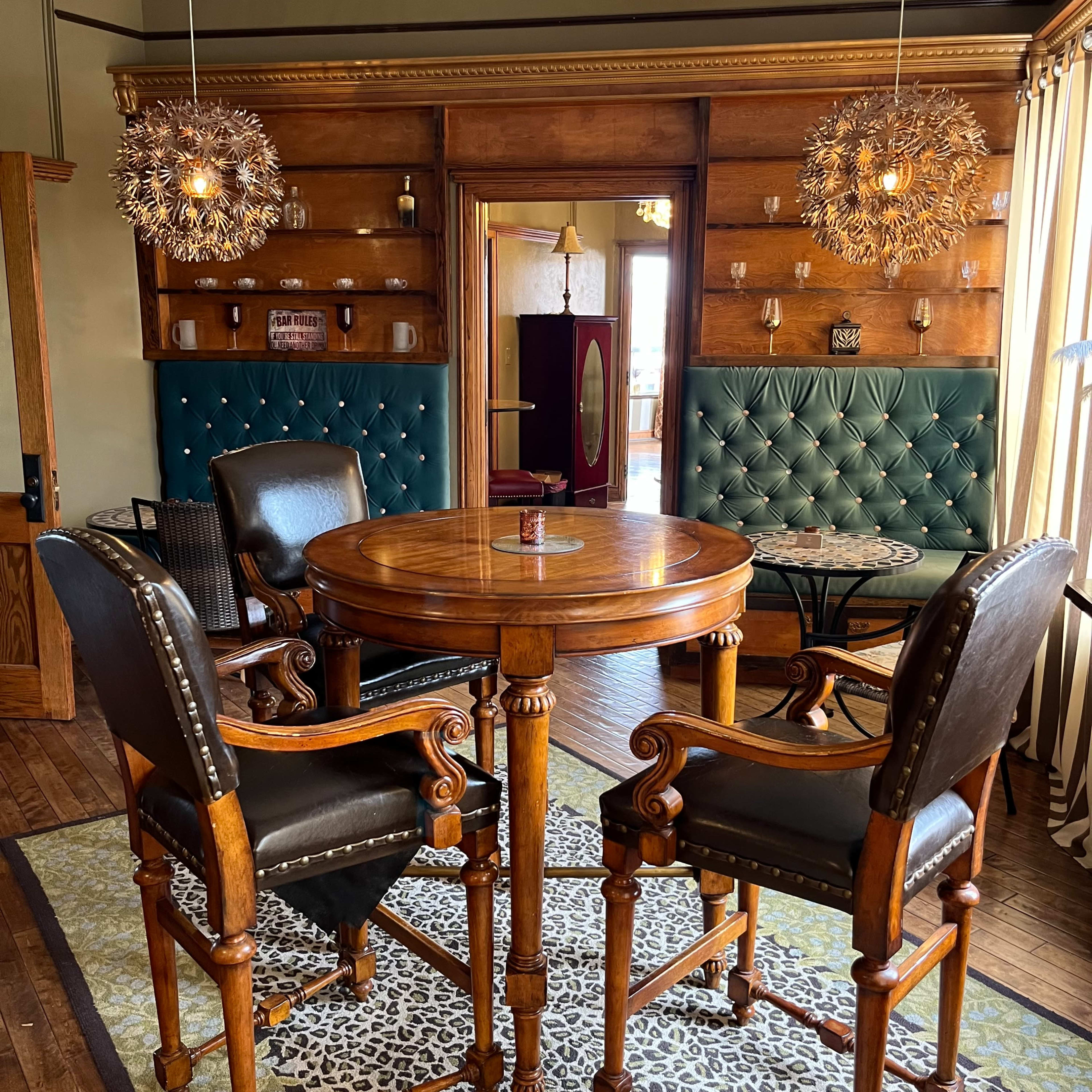 A restaurant interior featuring a round wooden table surrounded by two black leather chairs and a green upholstered wall with decorative lighting.