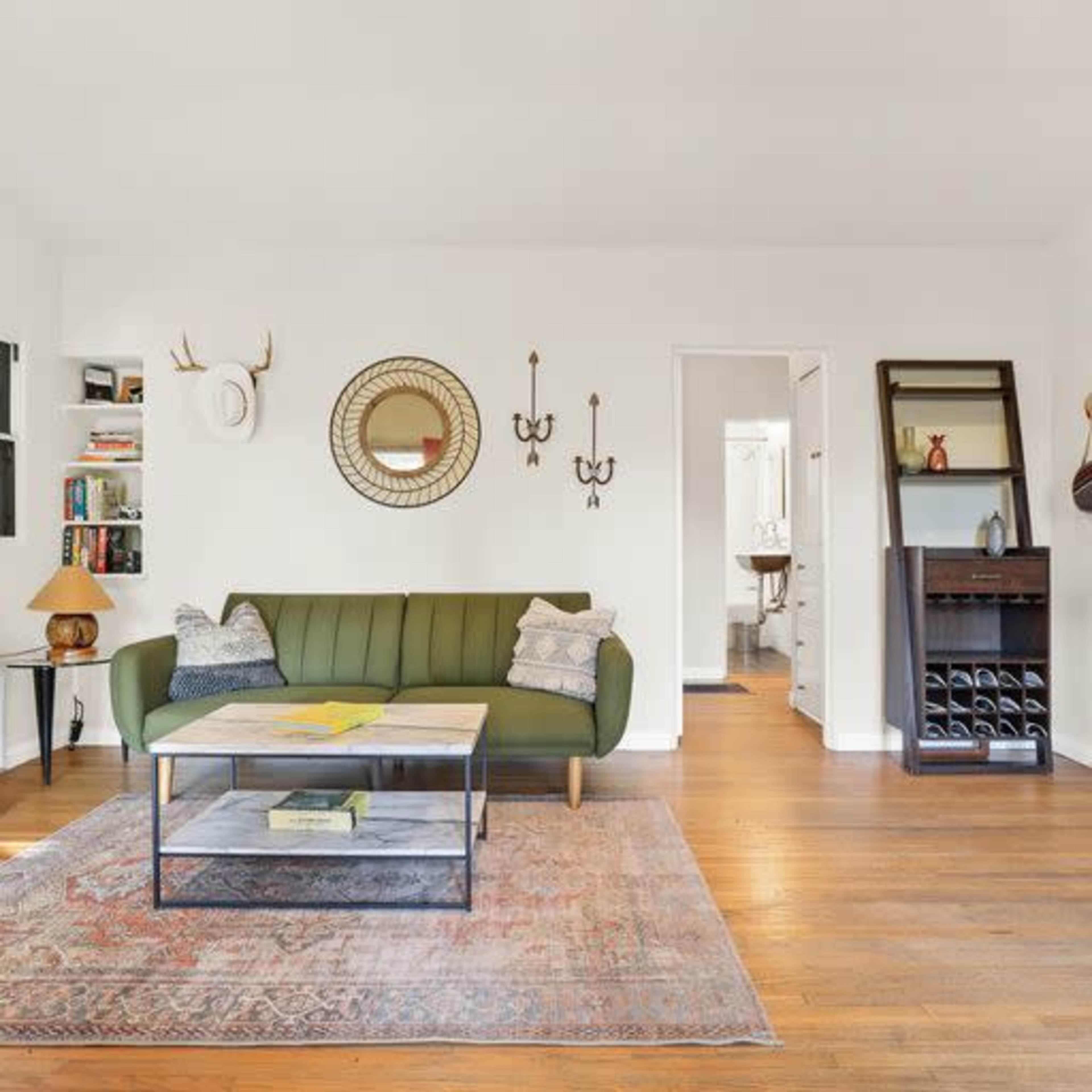 A living room with a green sofa, a coffee table, a round mirror on the wall, and access to a bathroom through an open door.