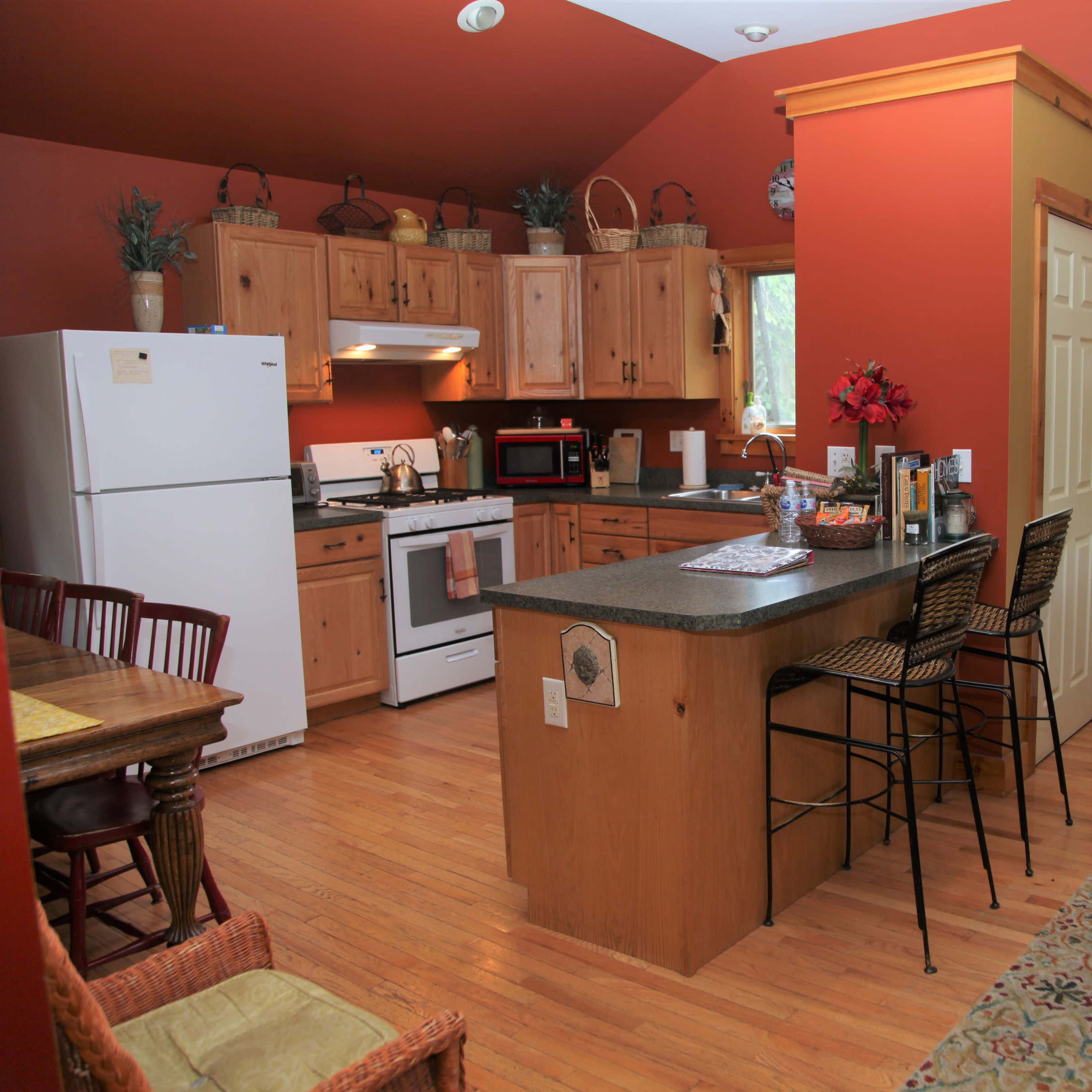 A cozy kitchen and living area with wooden cabinets, a white refrigerator, a dining table, and a red sofa, accented by warm wall colors.