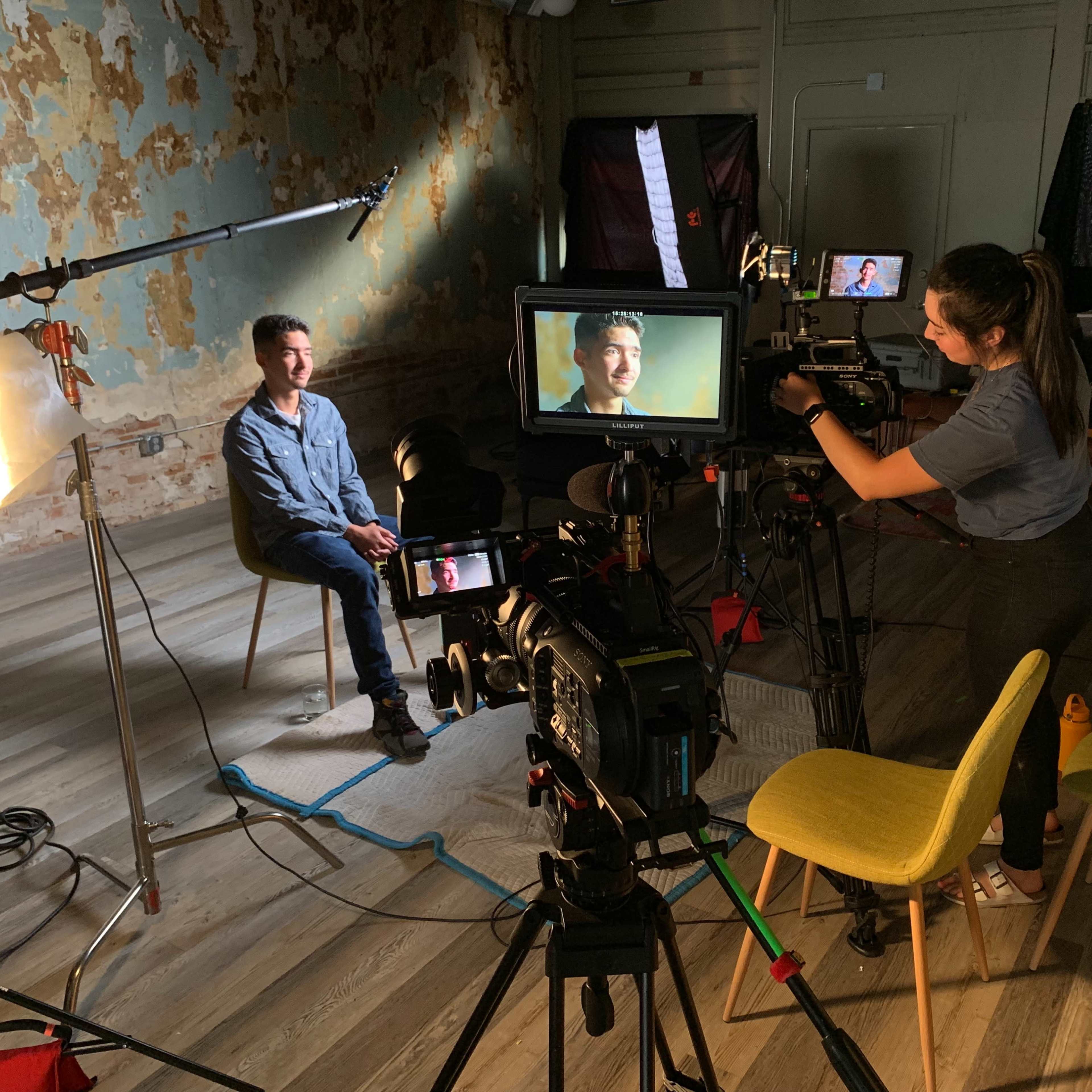 A person is being filmed in a seated interview setup, with cameras, lights, and a crew member adjusting equipment in a room with a distressed wall.