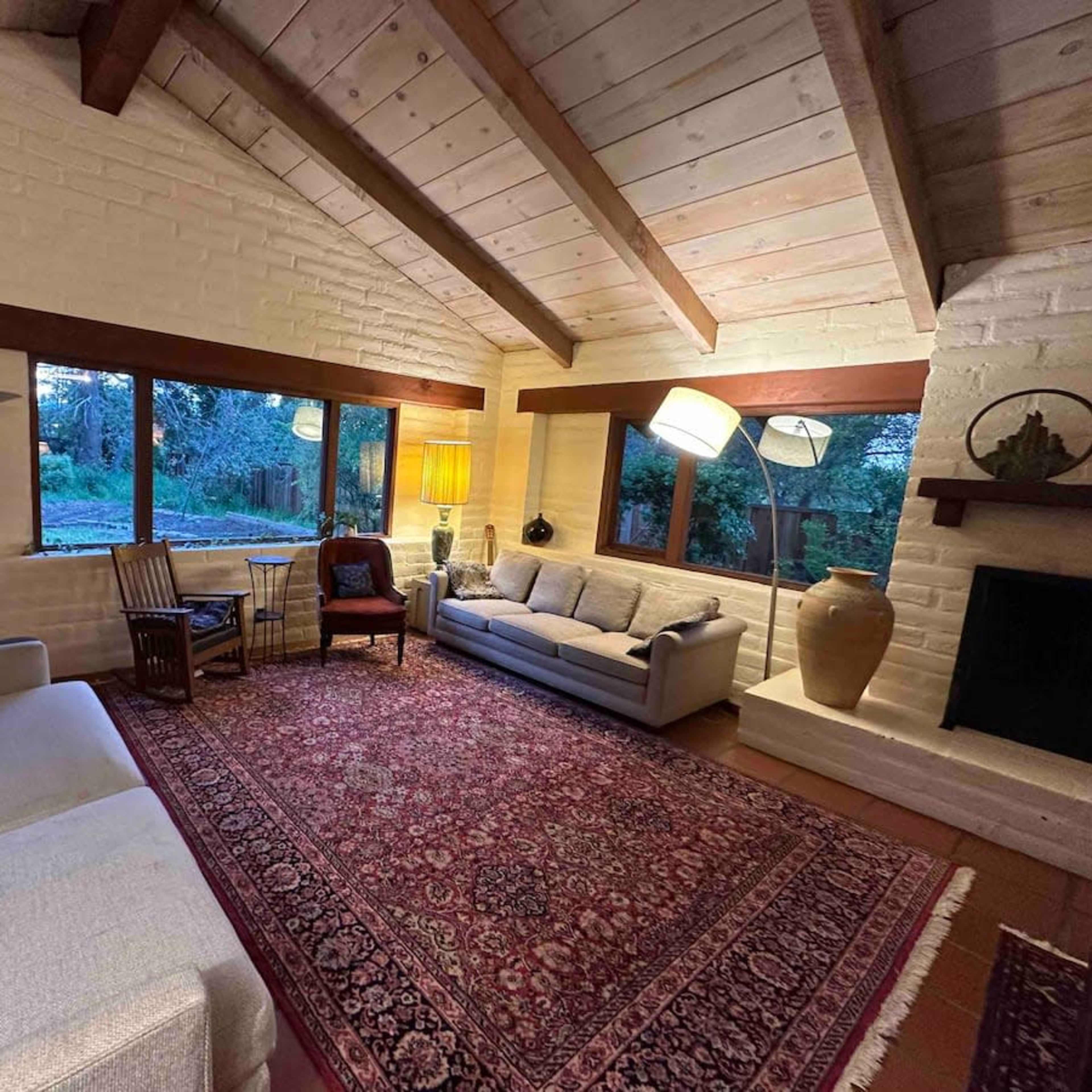 The scene displays a cozy living room featuring a gray sofa, wooden chairs, a large area rug, and large windows overlooking a garden.