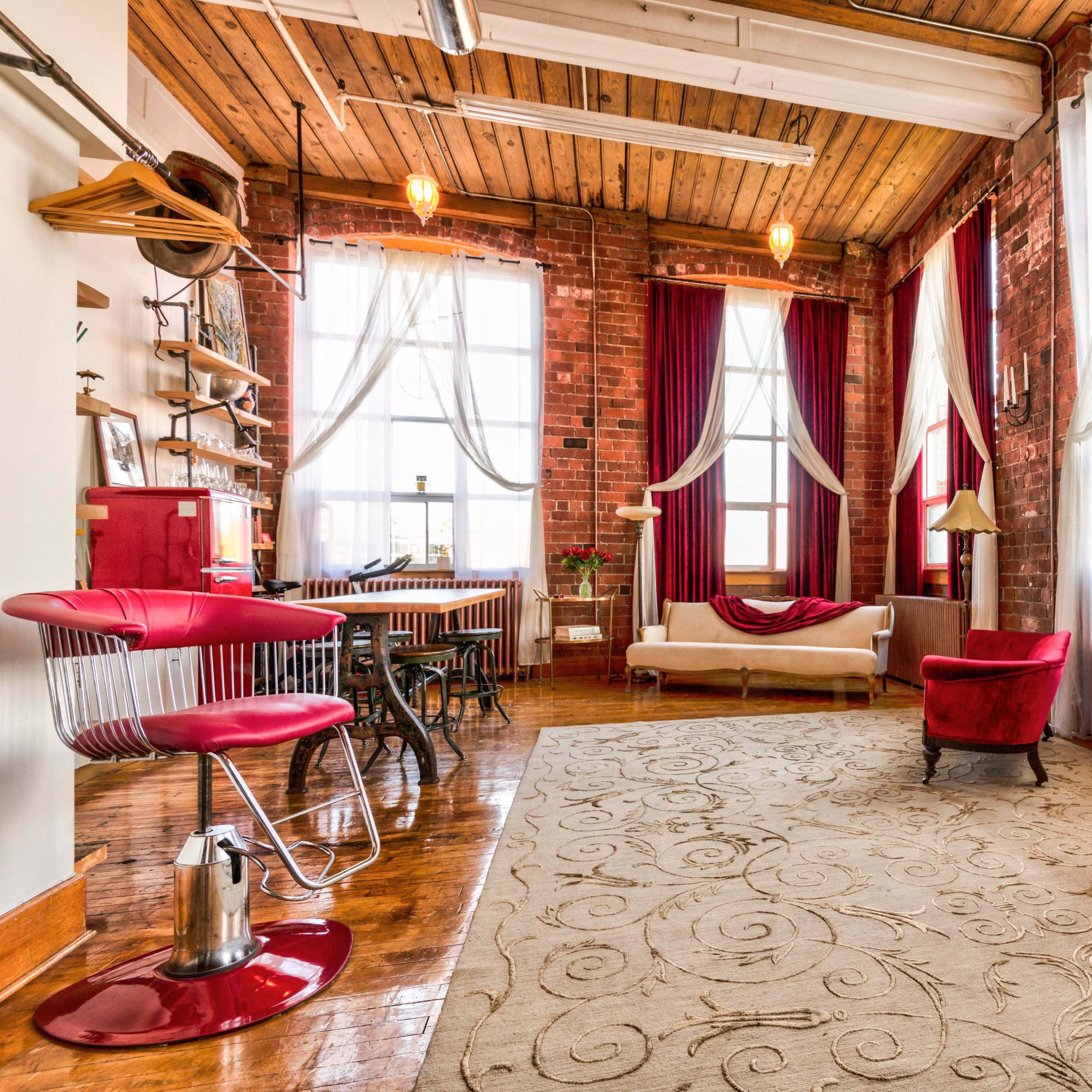 A spacious industrial-style room with exposed brick walls, large windows, and a mix of modern and vintage furniture.