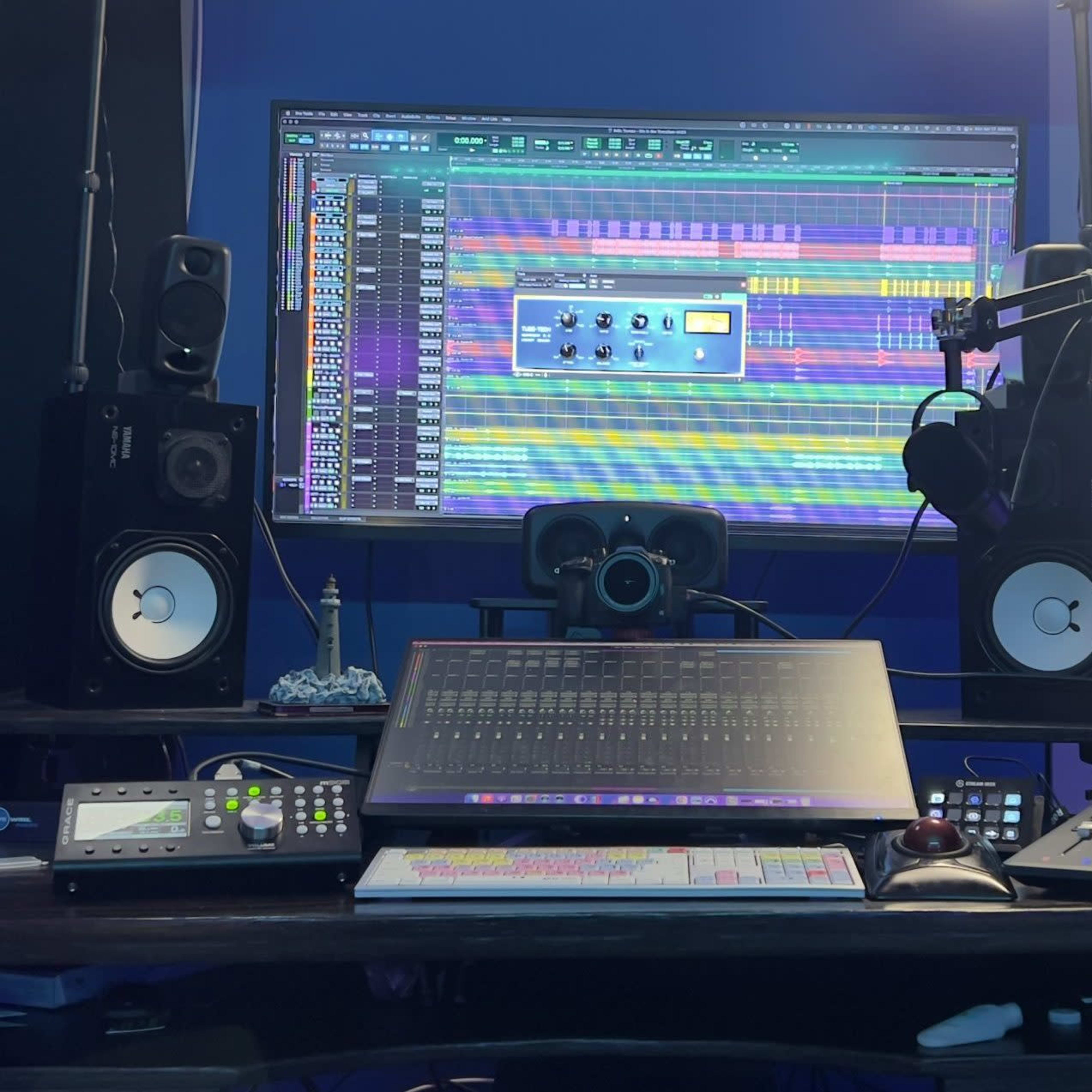 A music production studio setup with a large computer monitor displaying a digital audio workstation, flanked by speakers and various audio equipment on a desk.