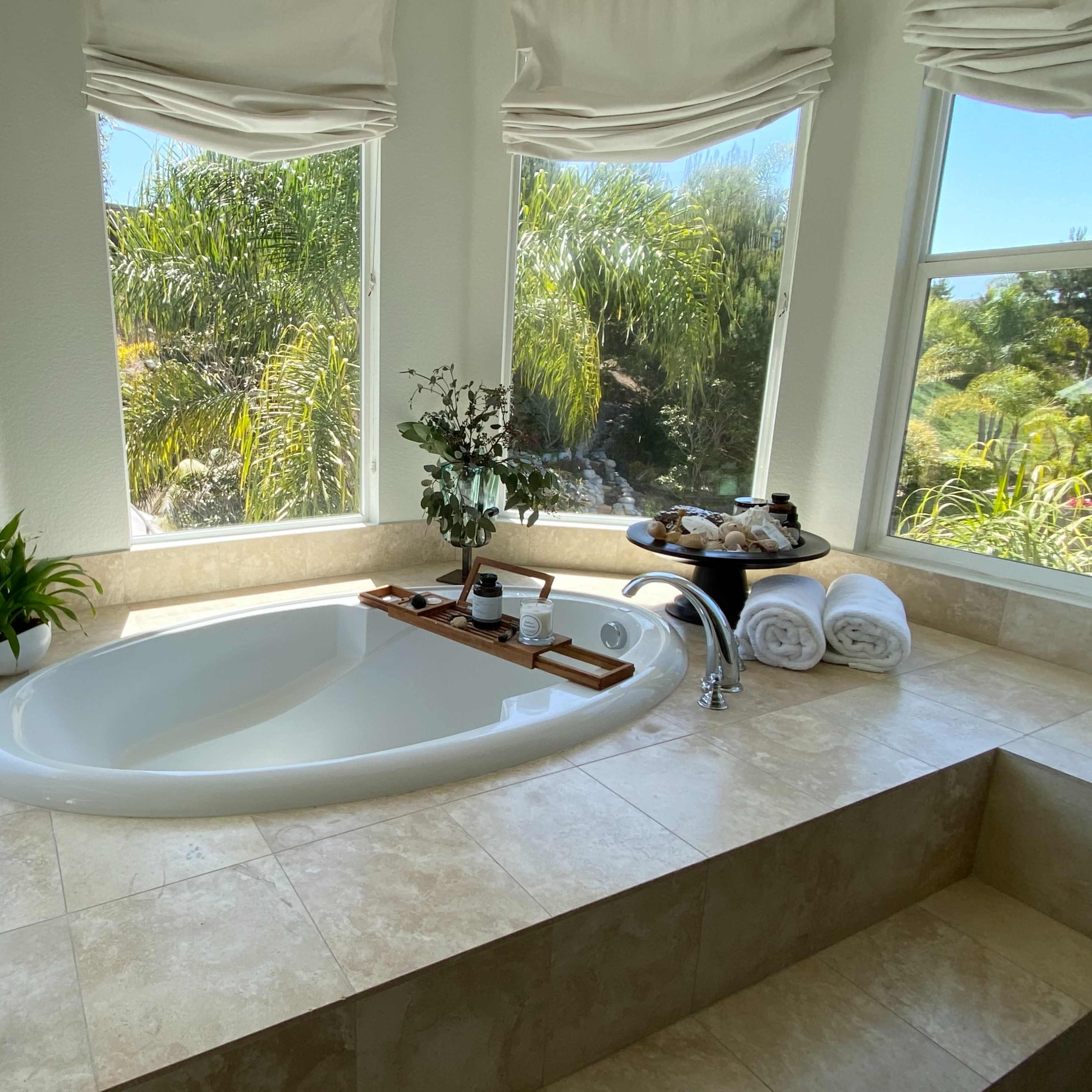 A large, oval bathtub is positioned by a window with views of greenery, accompanied by neatly arranged towels and decorations on a wooden tray.