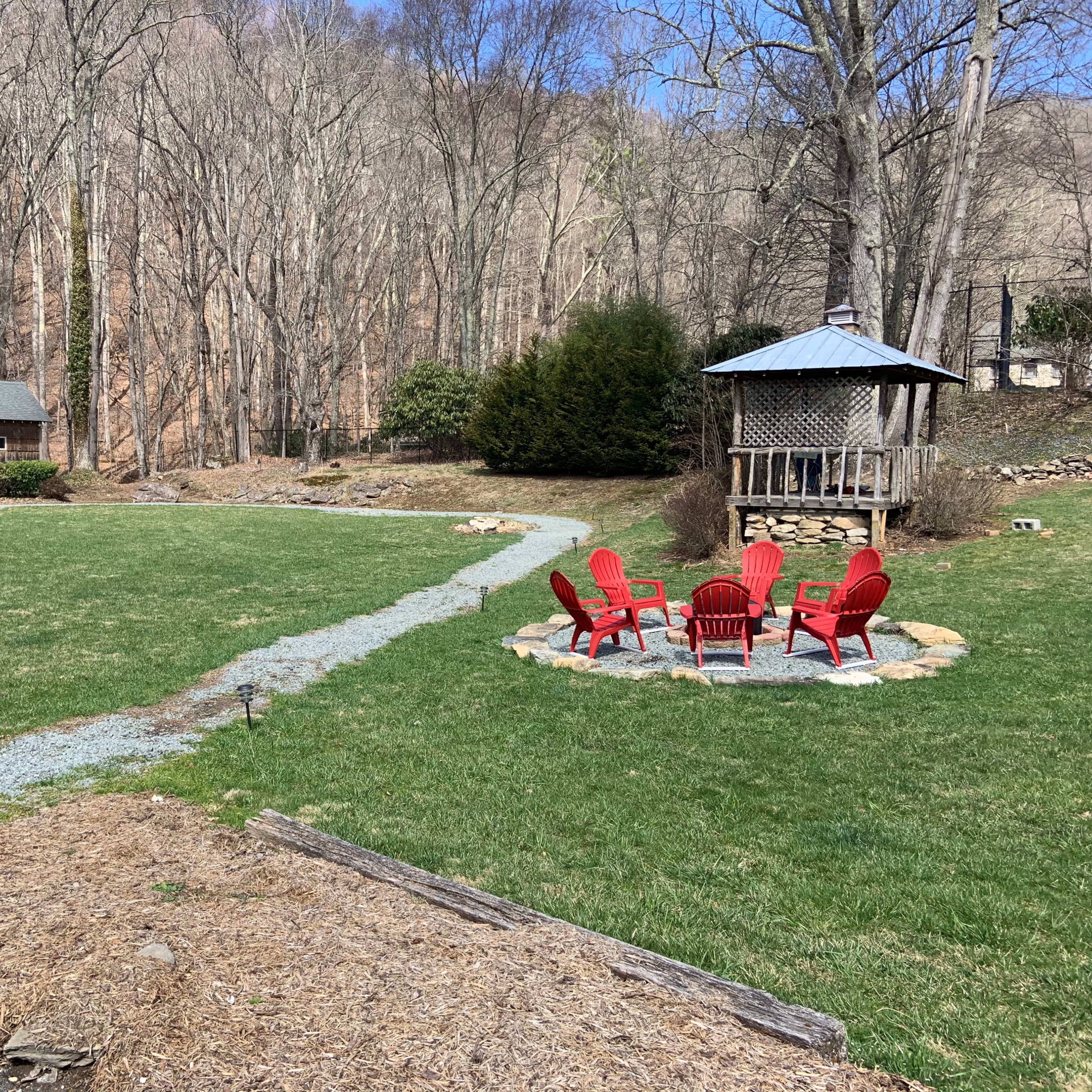 A grassy area with a stone path leading to a circular fire pit surrounded by red chairs, with a cabin in the background and trees beyond.