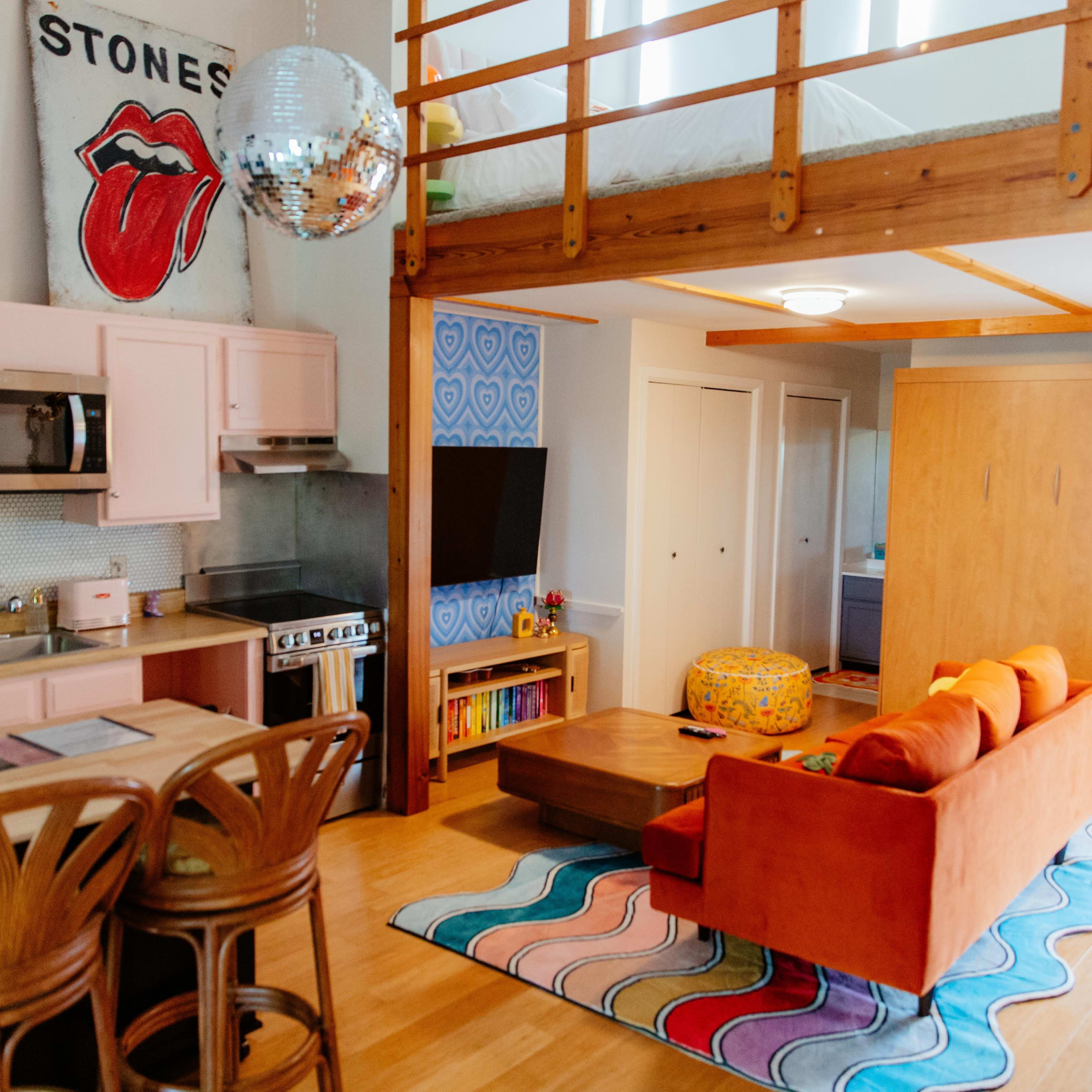 A modern open-plan living space with a kitchenette, a seating area featuring an orange sofa, and a lofted bed above, decorated with vibrant artwork and colorful furnishings.