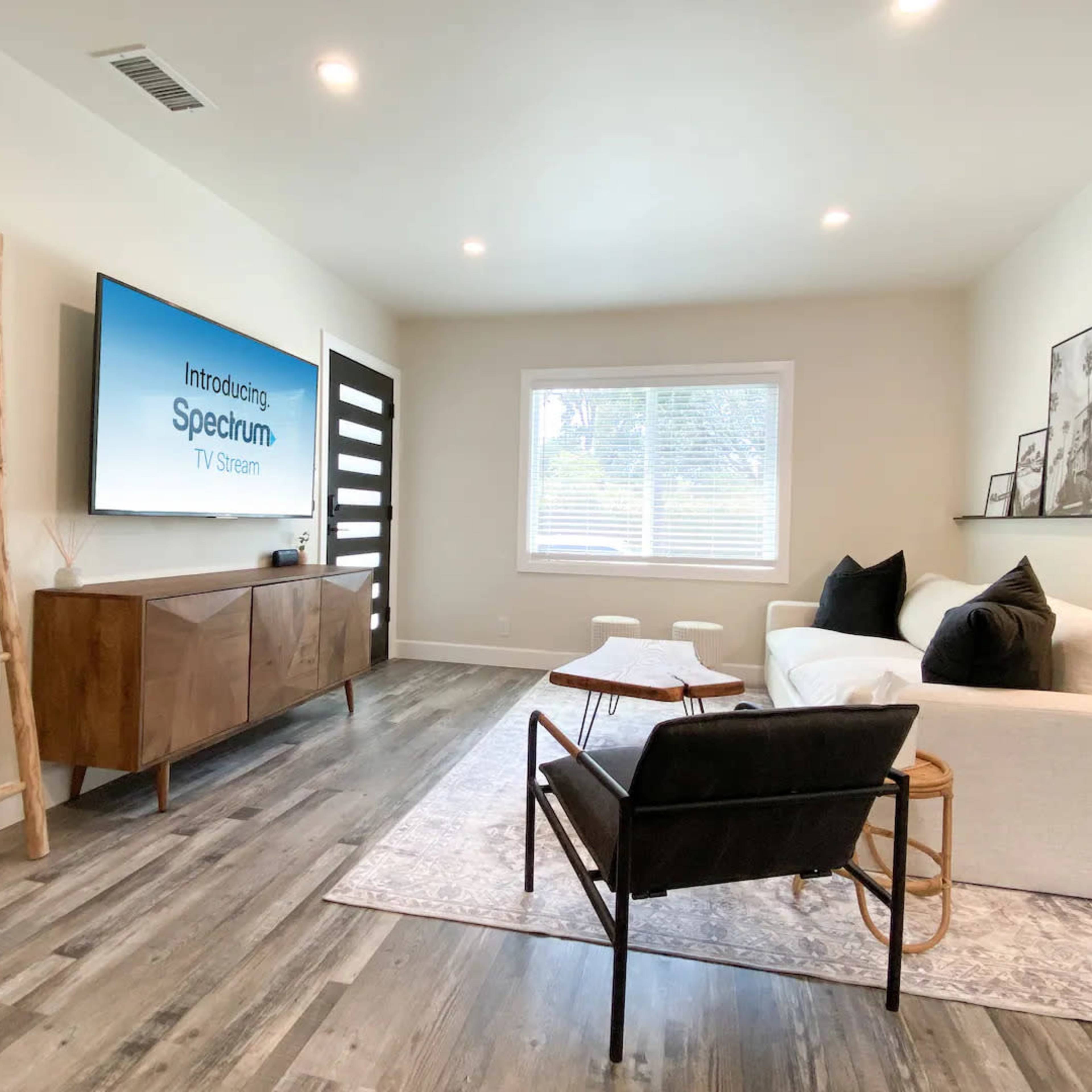 A modern living room features a white sofa, black chair, wooden media console, and a large television mounted on the wall.