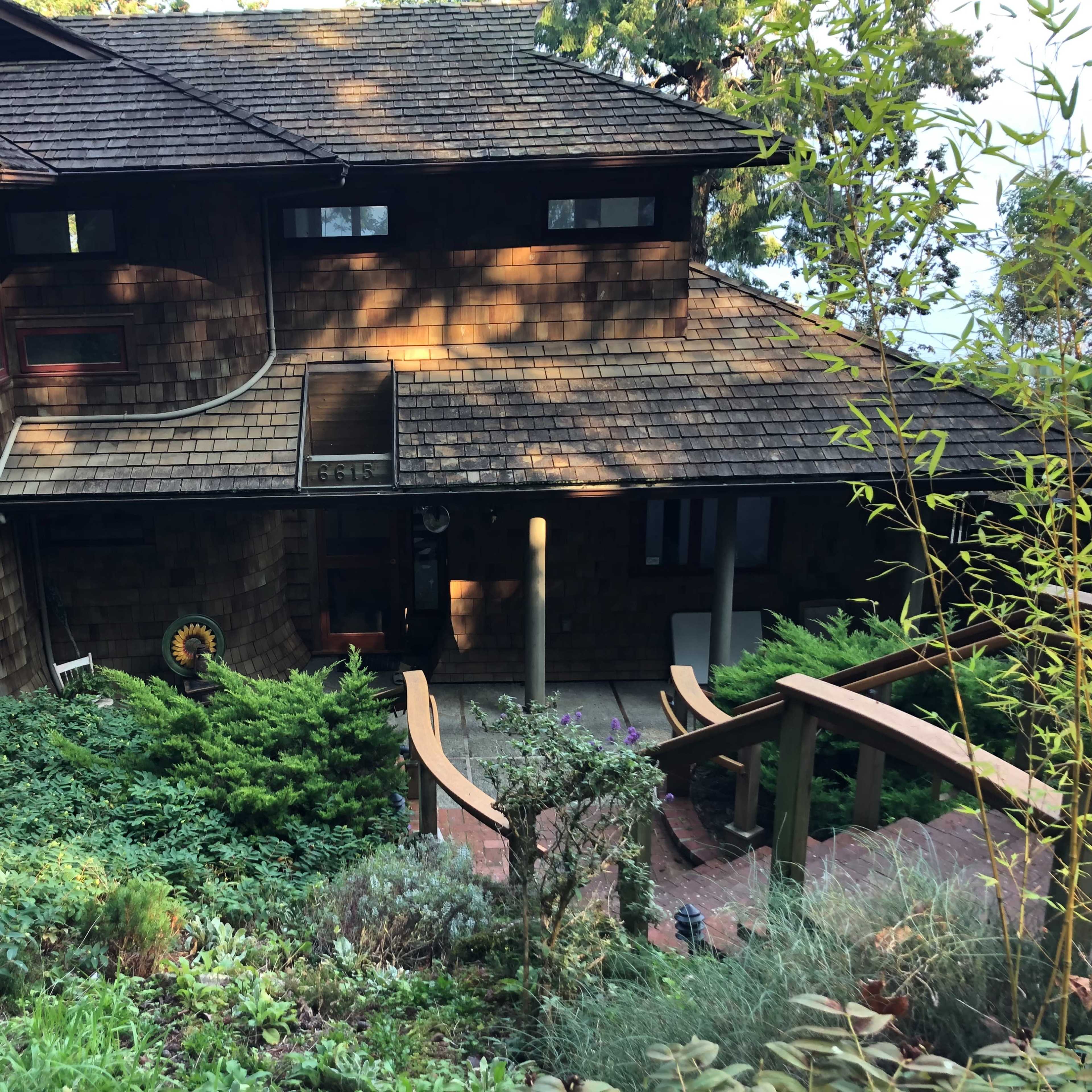 A large, multi-level wooden house with a steeply sloped roof is set among lush greenery and steps leading to its entrance.