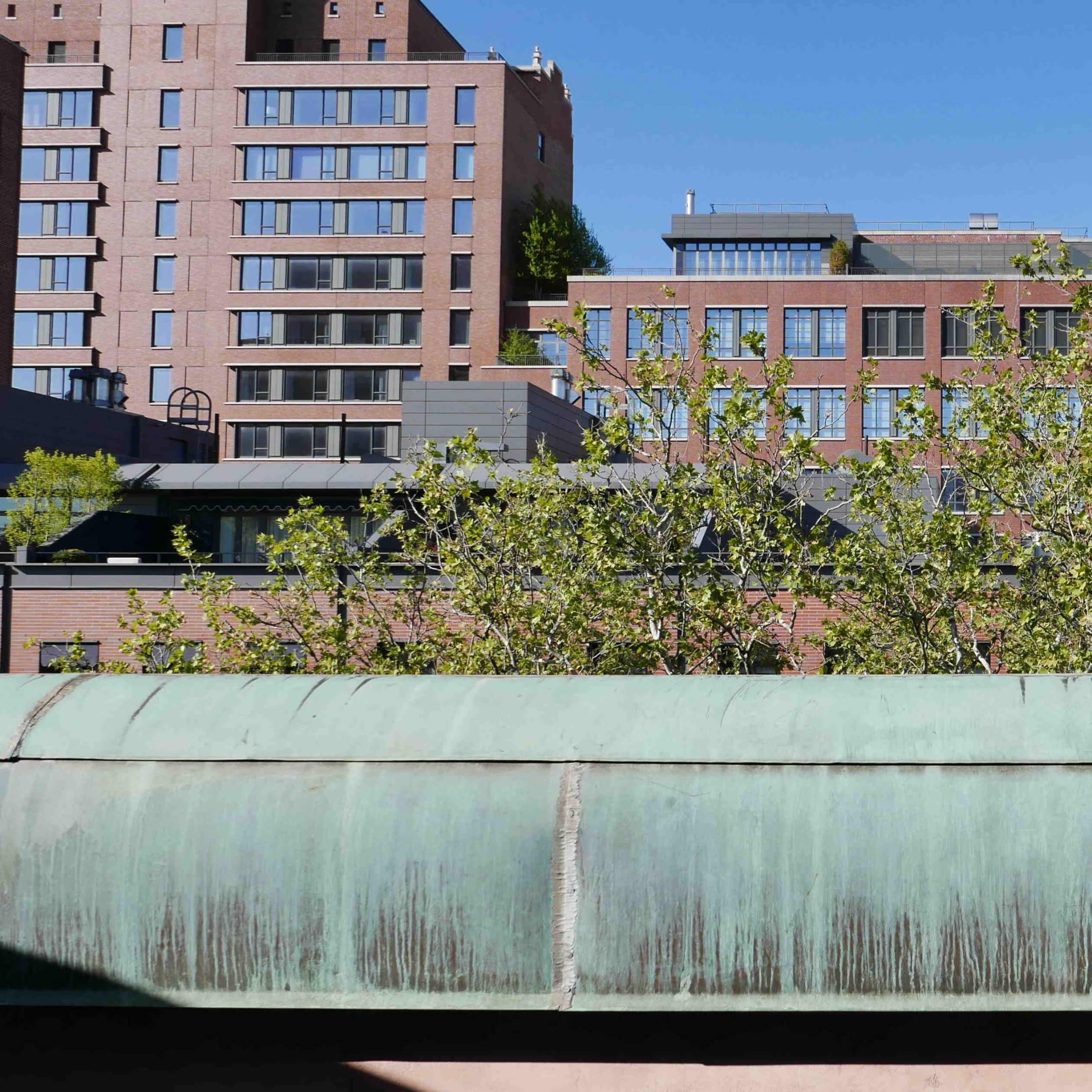 A view of brick and modern buildings lining a street, with greenery in the foreground and a copper-colored structure below.