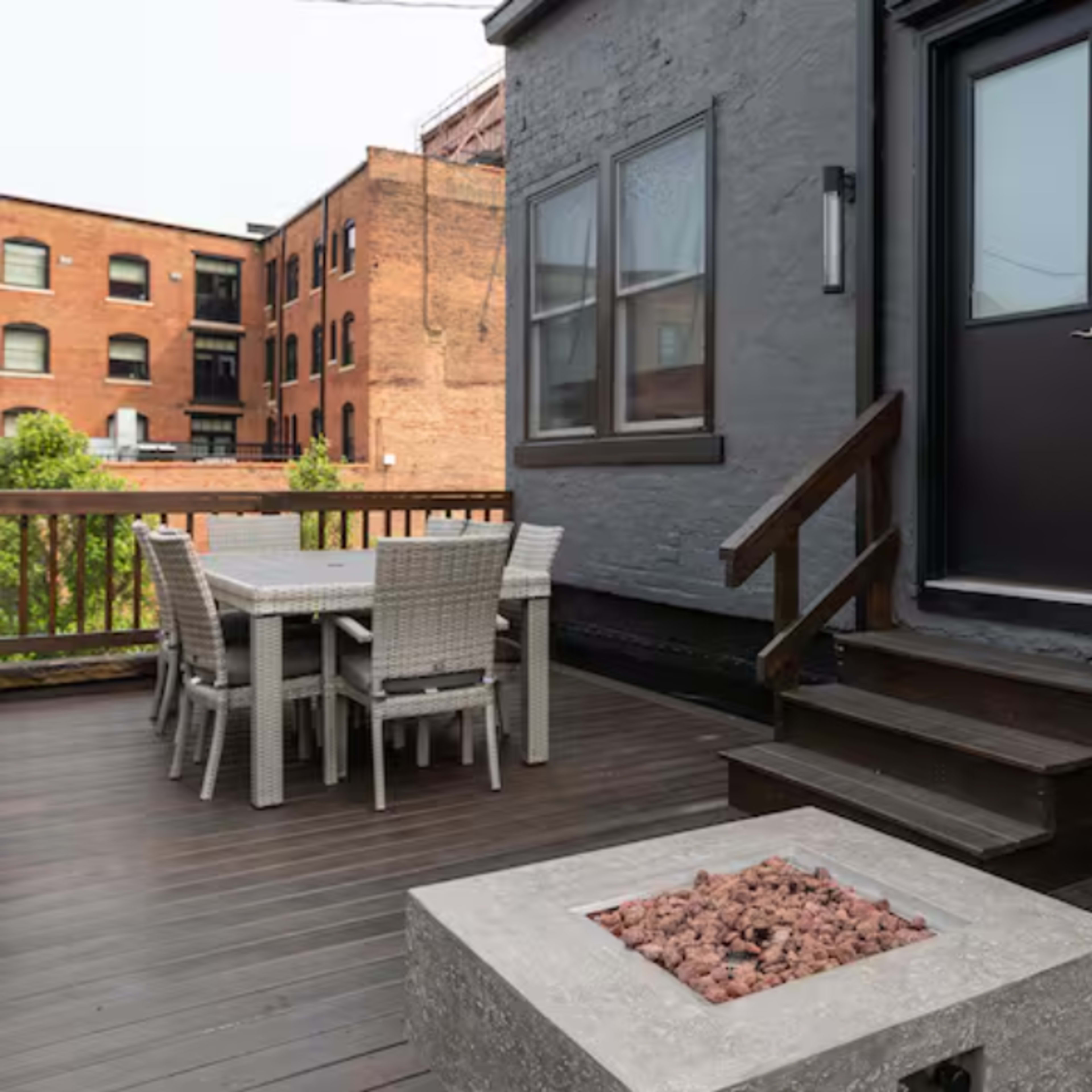 A modern deck with a seating area, a dining table, and a fire pit, adjacent to a black door and surrounded by brick buildings.